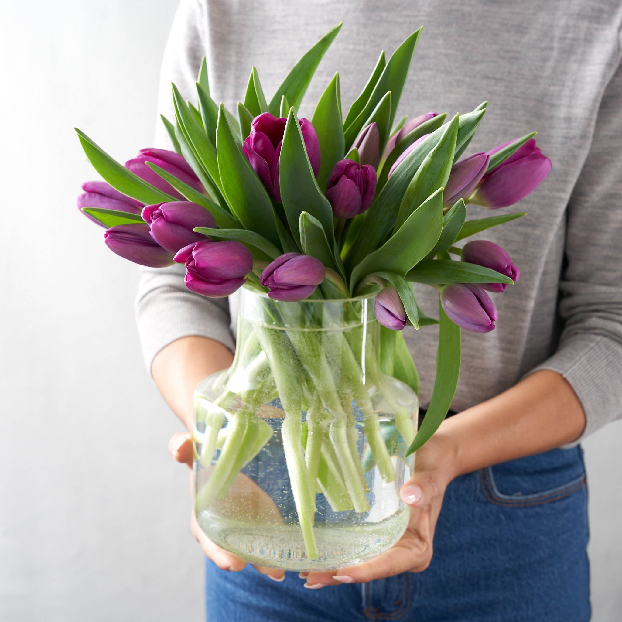 Woman in grey sweater holding a rounded clear glass vase filled with purple tulips