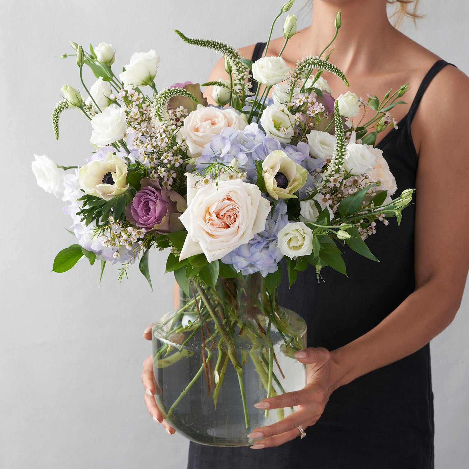 Woman in sleaveless black dress holding glass vase full of blue cream lavender and white flowers.
