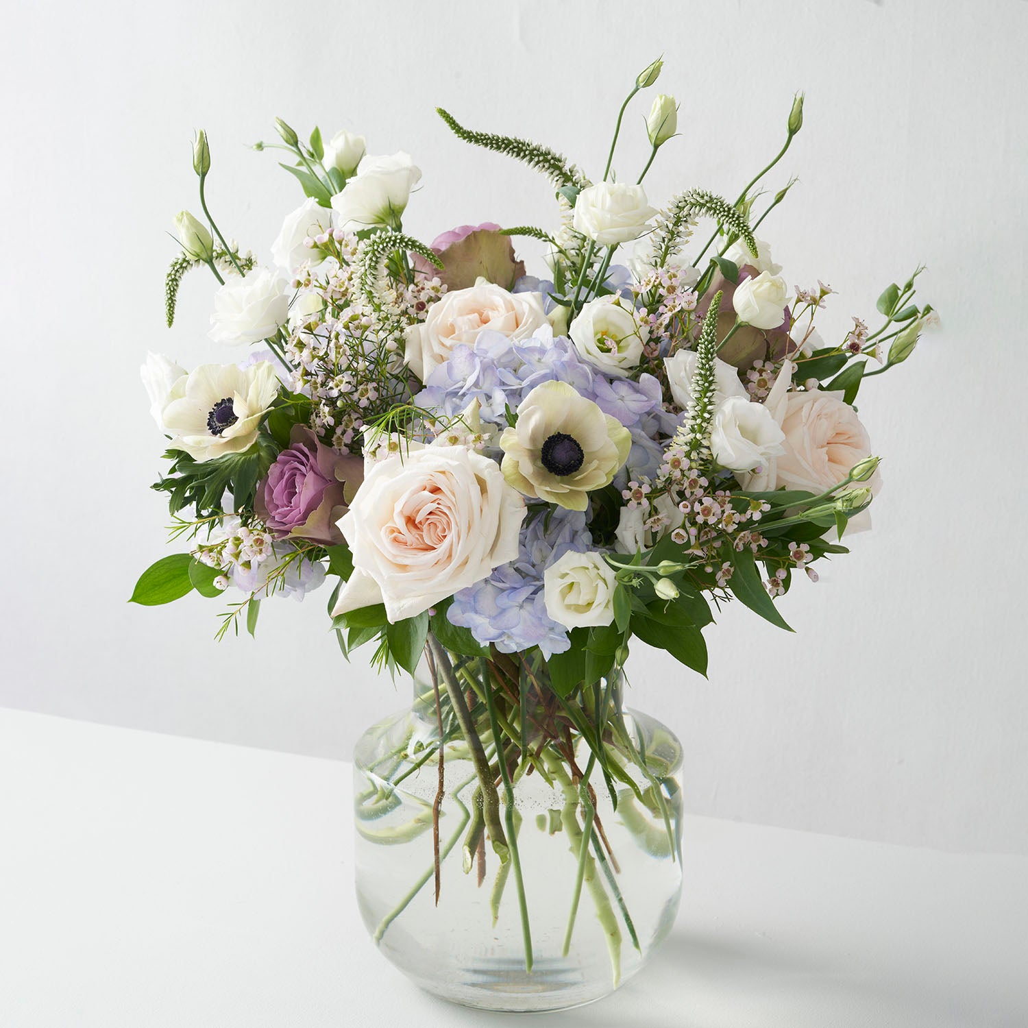 Glass vase full of blue, lavender, cream and white flowers on white background.