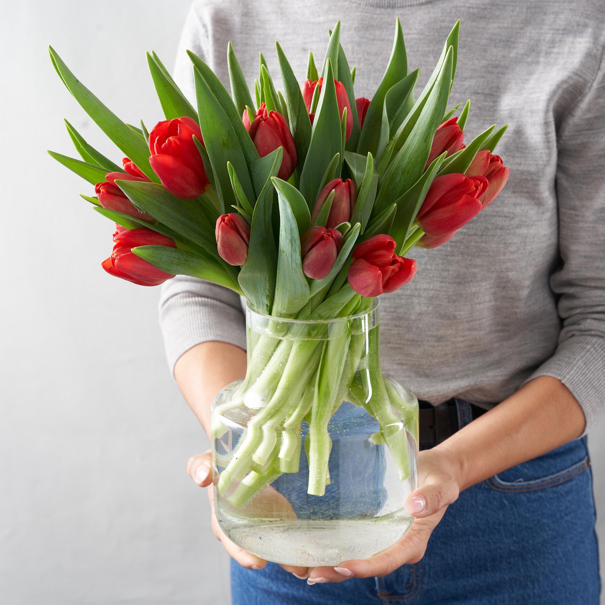 Woman in grey shirt holding a clear glass vase filled with a dozen red tulips