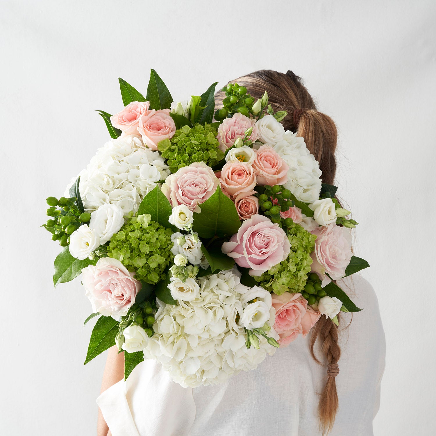 A view fromthe back of a woman in white shirt with long braid holding large round pink white and green bouqet of roses and hydrangea.