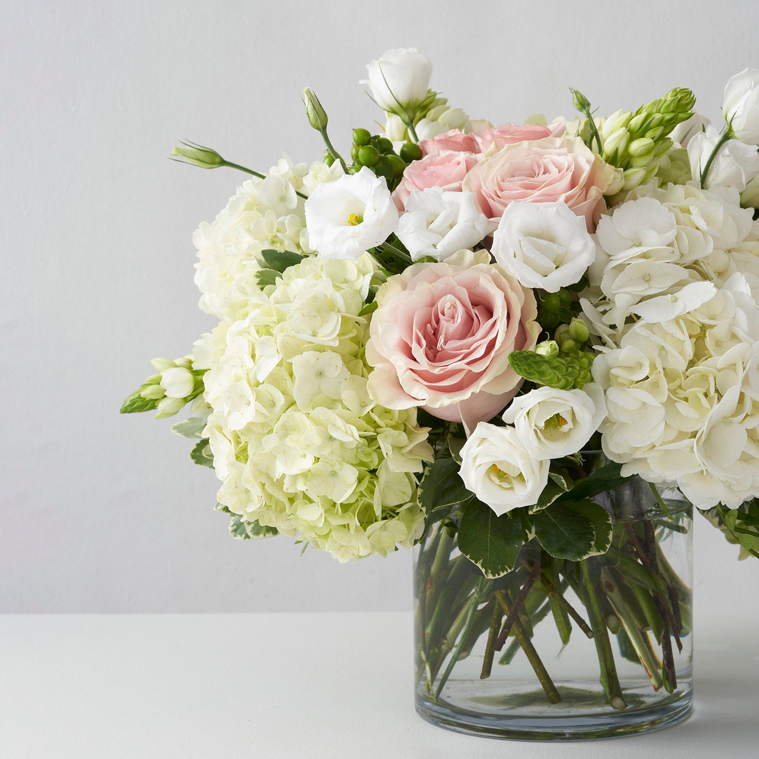 Pink Mondial roses, white hydrangea, and white lisianthus in clear glass vase on white backfground.