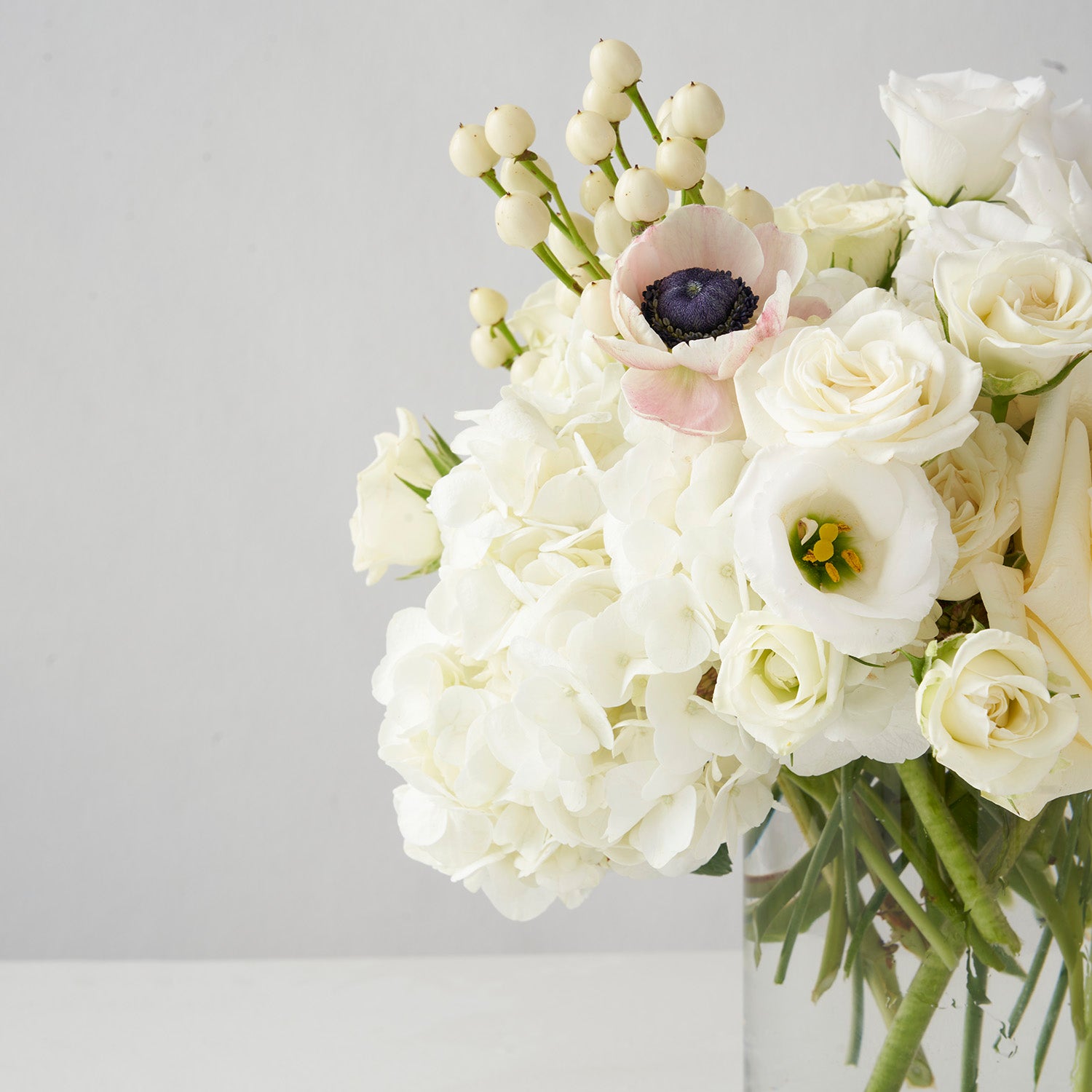 Off center image of clear glass vase filled with white flowers including roses, anemonies, and lisianthus and stems in water on white background.