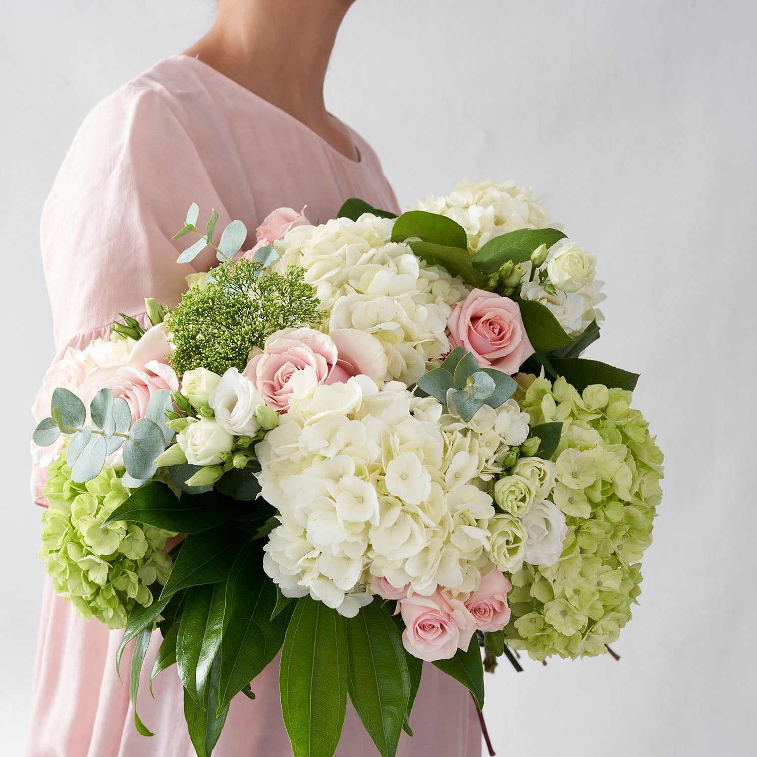 Woman in pink dress, holding bouquet of white and green  hydrangea, soft pink roses and eucalyptus.