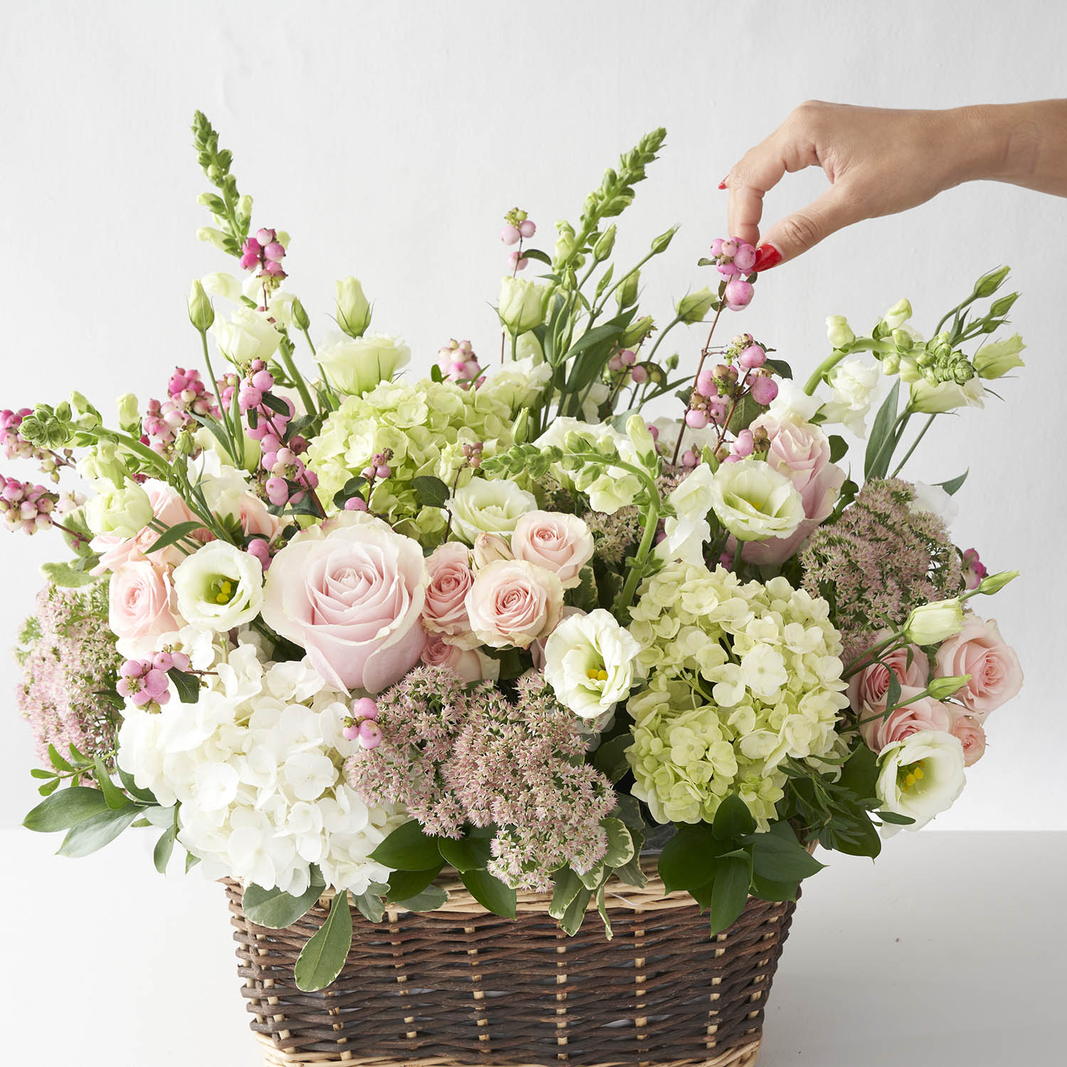 Woman's hand arranging pale pink, soft green and white flowers in a natural basket.