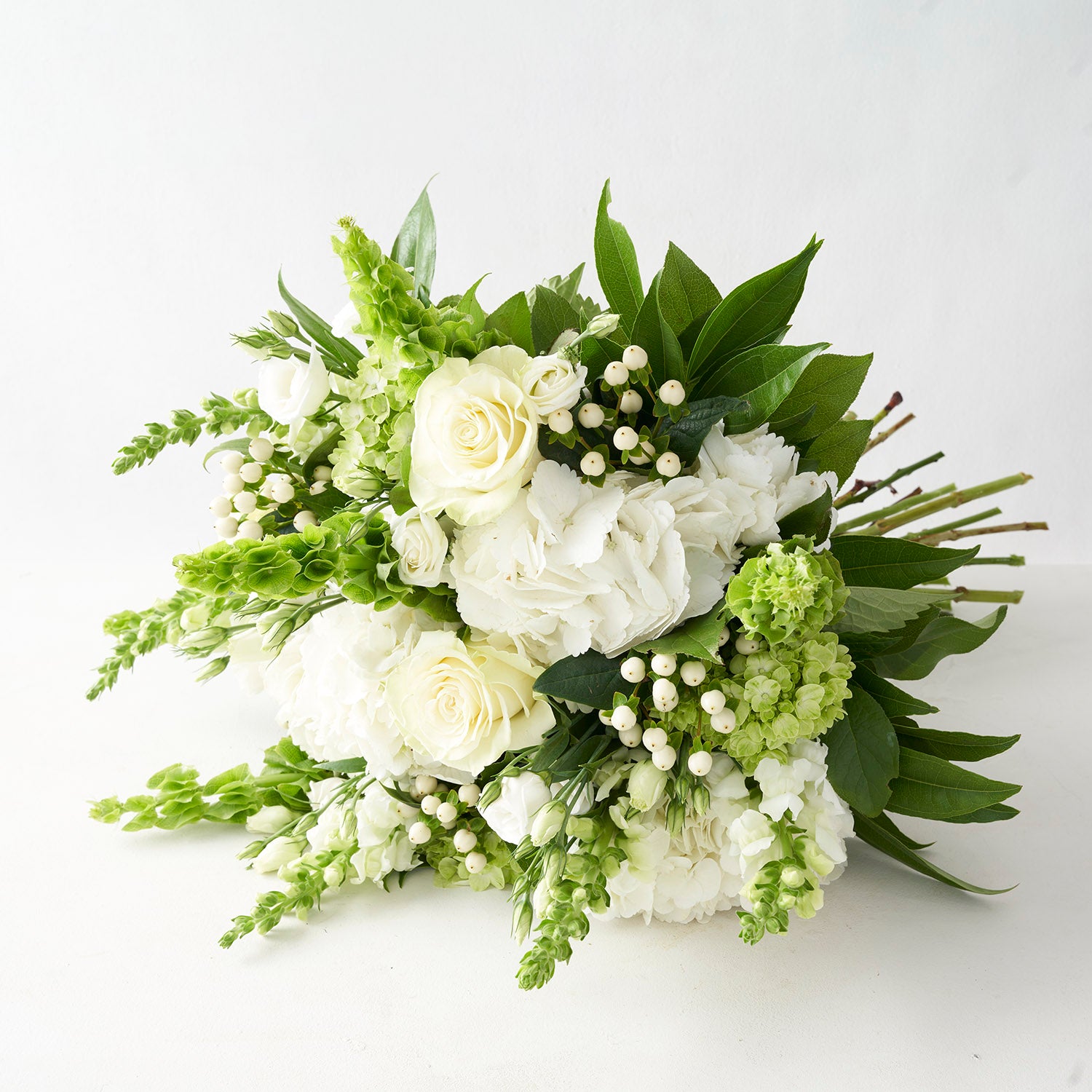 A round white and green bouquet including white roses, white and green hydrangea, and white snapdragons  laying on white background.