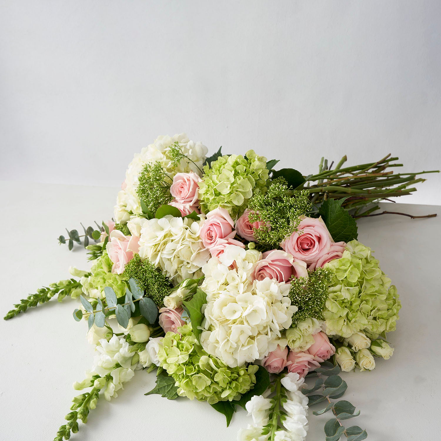 Bouquet of pale green white and pale pink flowers, uncluding hydrangea, roses, snap dragons, and eucalyptus, laid out on white surface. 