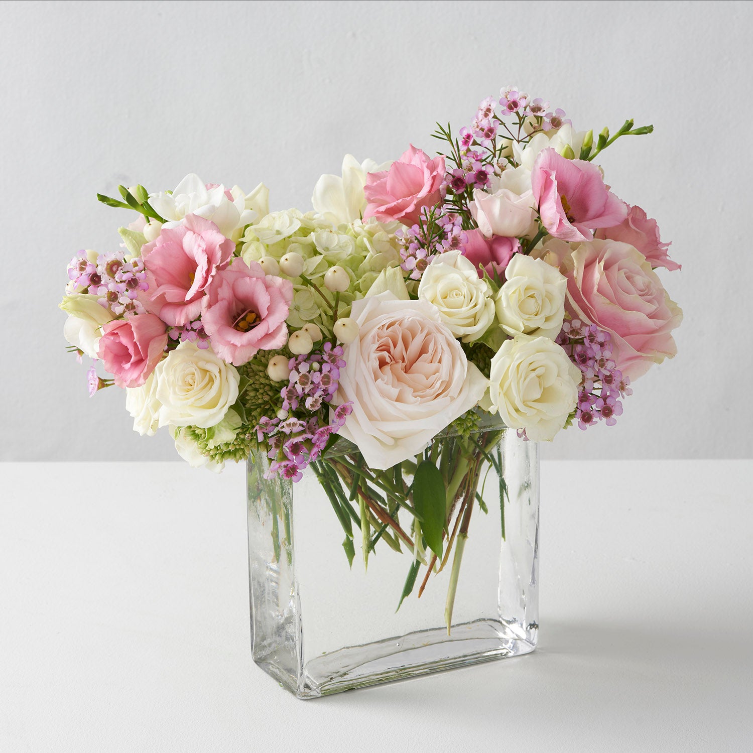 glass rectangular vase filled with O'hara roses pink Mondial roses white spray roses freesia and hydrangea on white background.