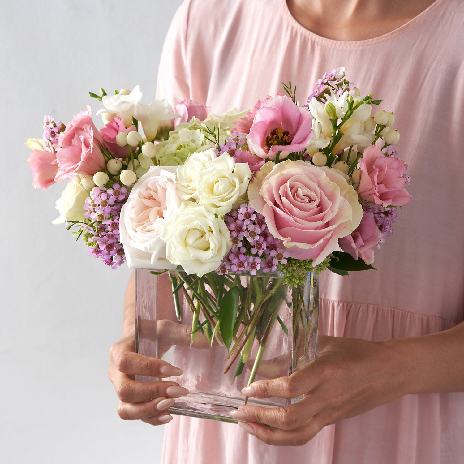 Woman in pink dress holding clearglass vase ful of variouse pink and white flowers.