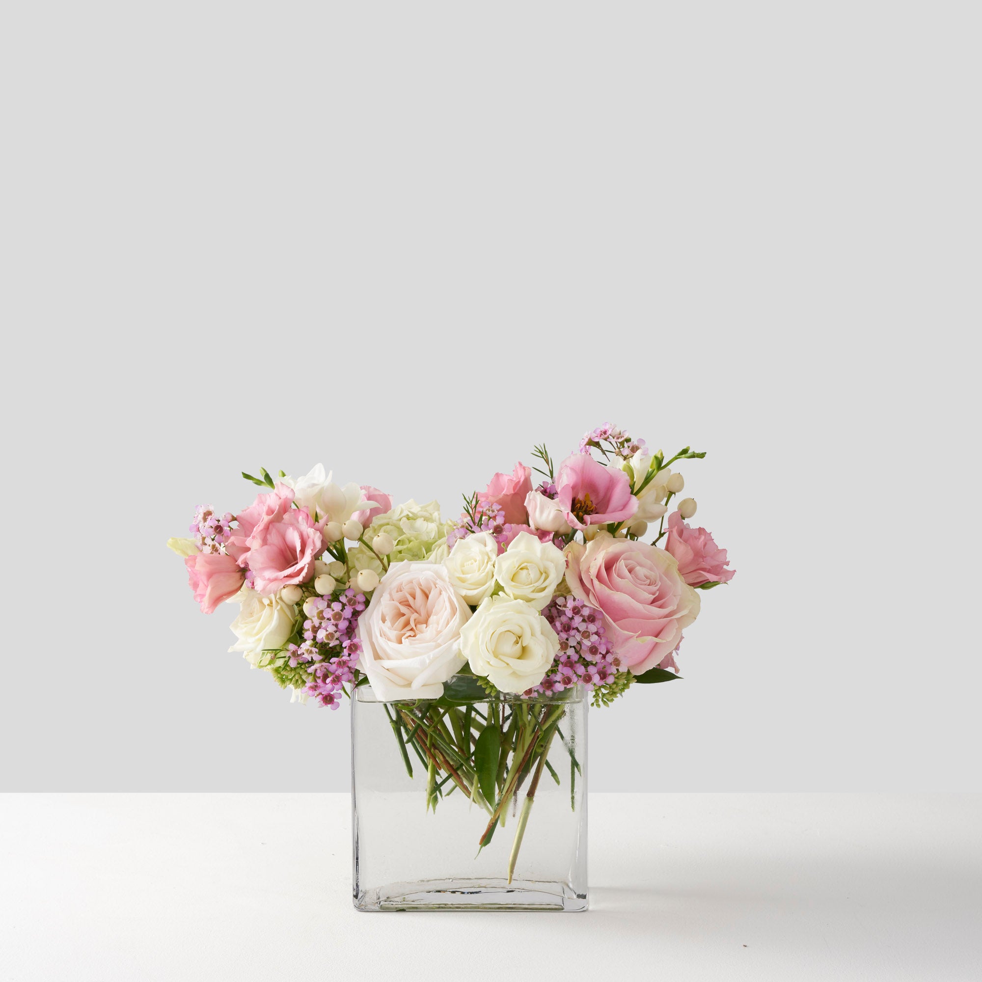 Pale pink and cream roses, white hydrangea, pink wax, and pink lisianthus flowers, arranged in a rectangular glass vase on white background.