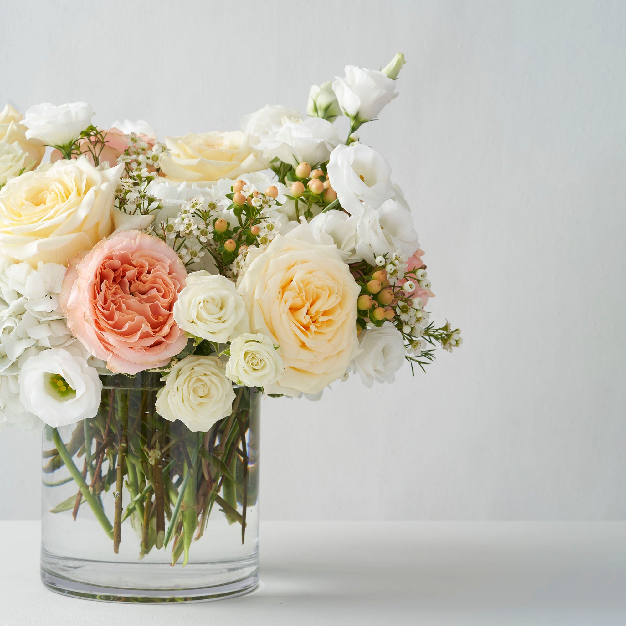 Peach roses, cream roses, white lisianthus, and white hydrangea arranged in glass vase on white background.