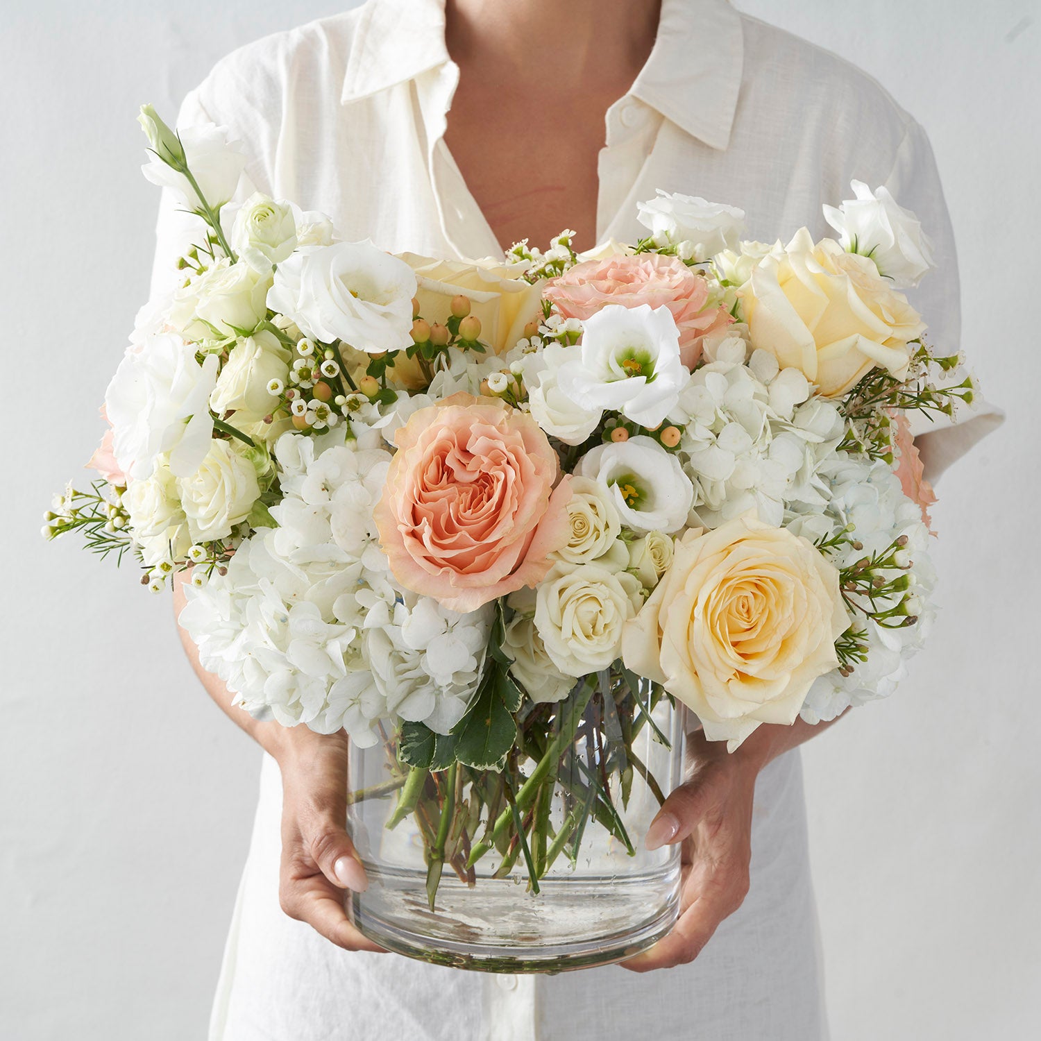 woman wearing all whit holding large clear glass vase filled with peach and cream roses,  white hydrangea and white lisianthus