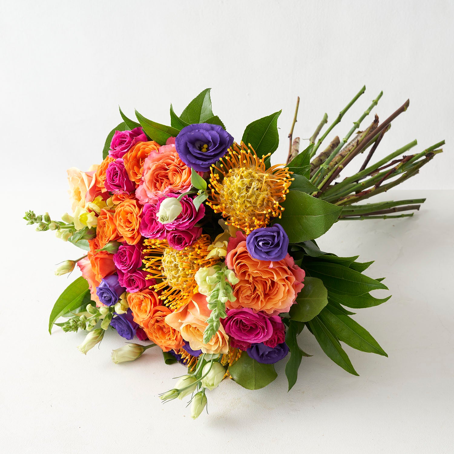 Round handtied bouquet with orange free spirit roses, dark purple lisianthus, hot pink spray roses, and gold protea on white background.