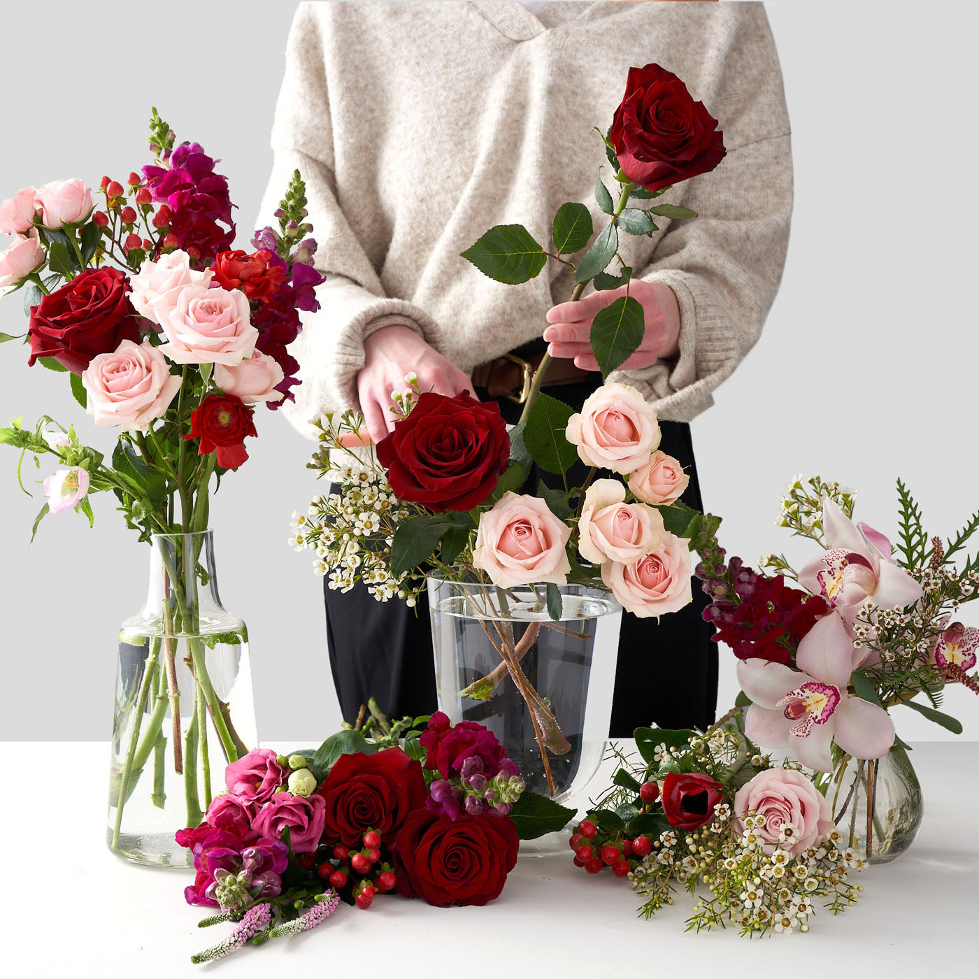 Woman in grey sweater arranging light pink spray roses, red roses, baby's breath, red hypericum berries, and purple veronica's inside of clear glass vases