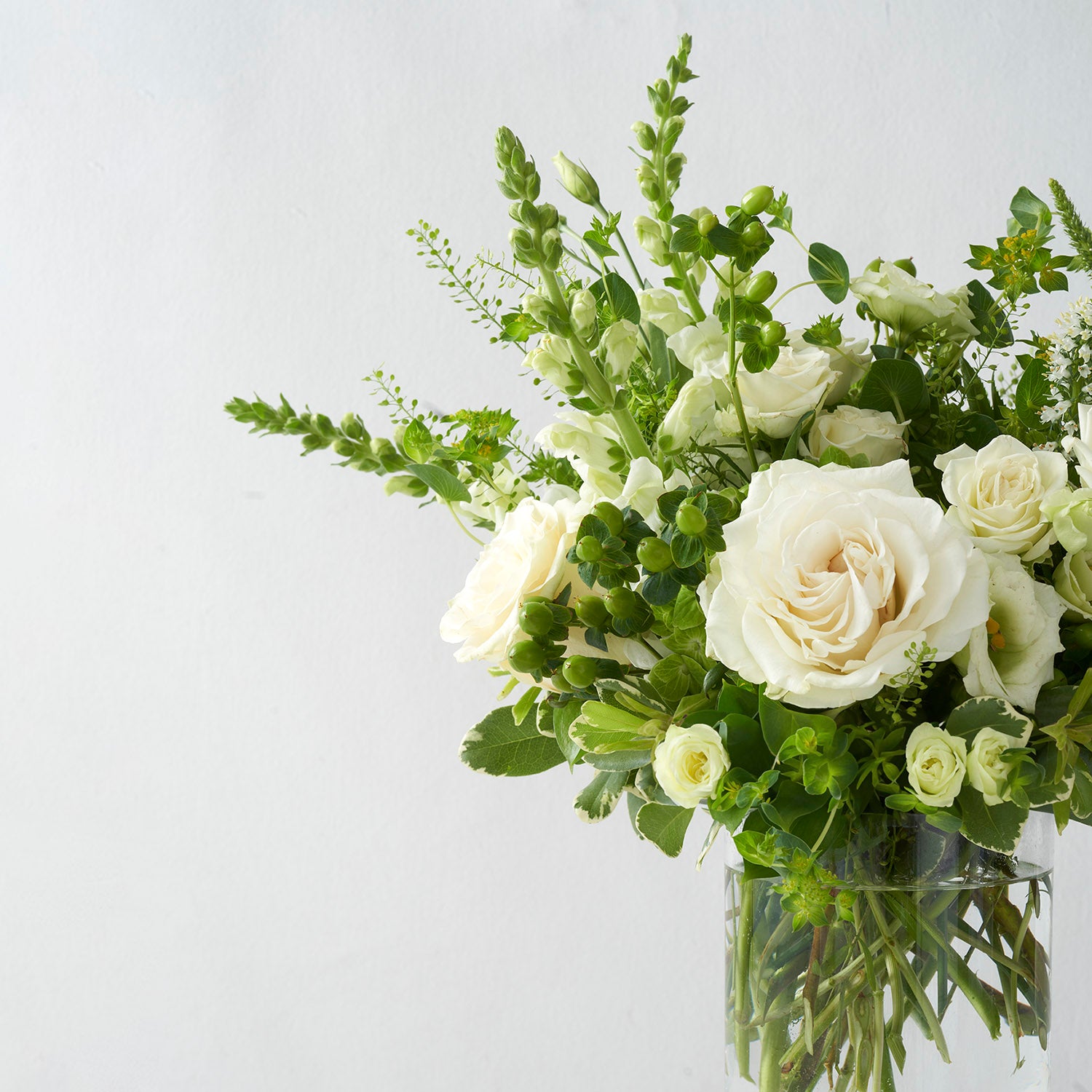 Arrangement of all white and green flowers, including roses lisianthus ans snapdragons, in glass vase on white background.