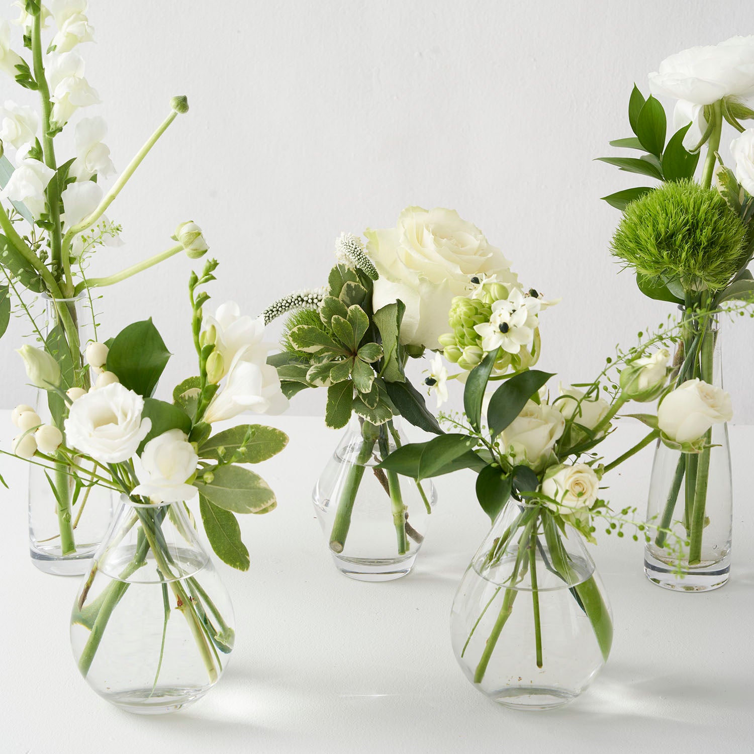 Five small glass vases filled with small white and green flowers including roses and snap dragons on white background