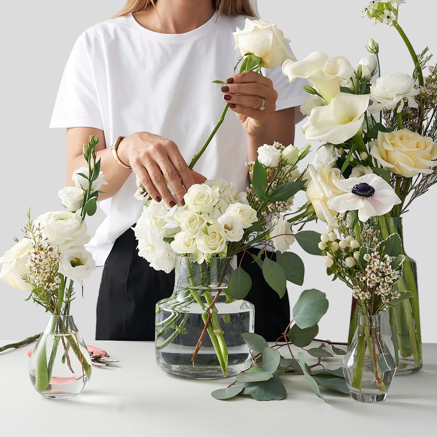 Woman in white shirt placing white rose into glass vase surrounded by other white flowers.