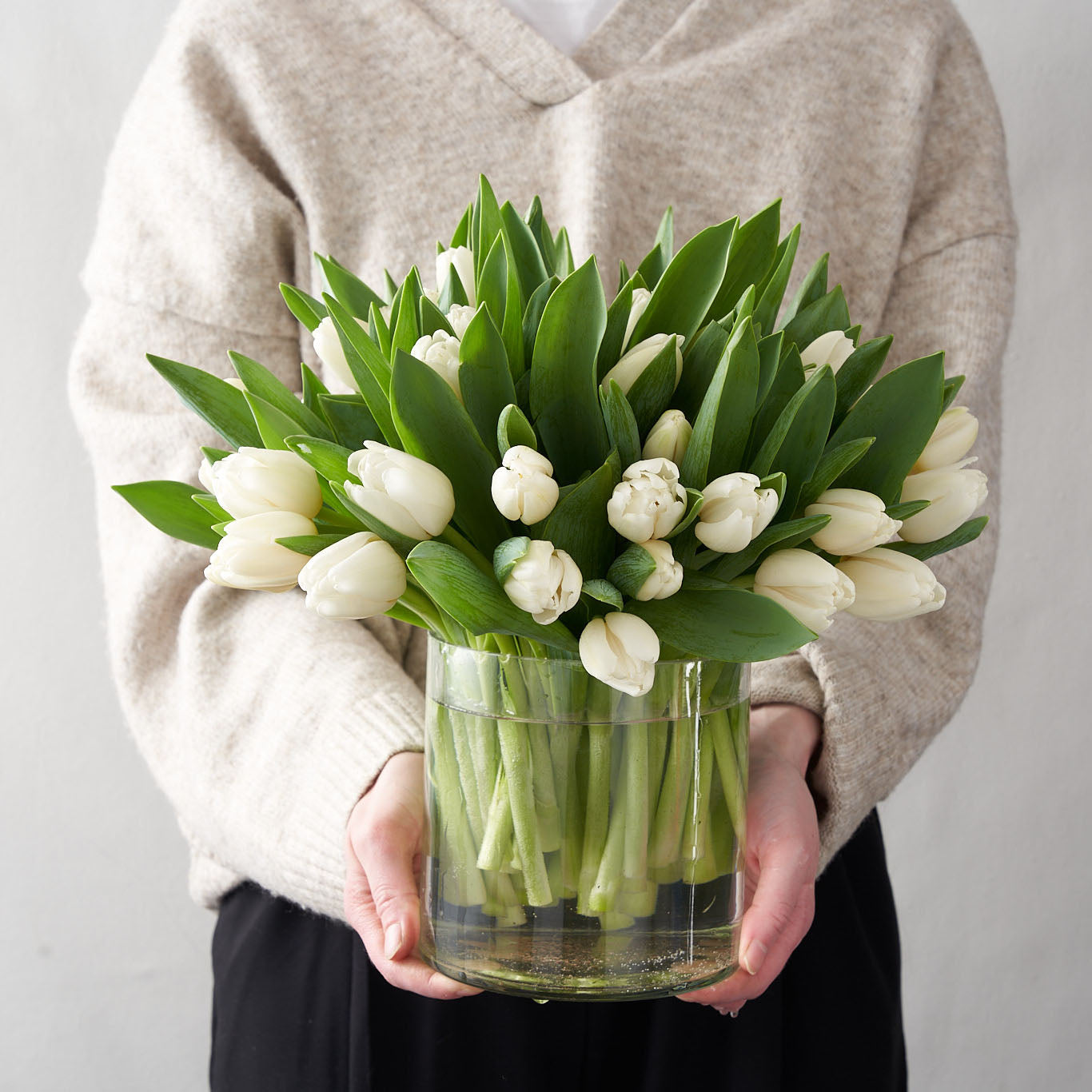 Woman in grey sweater holding a cylindrical clear glass vase containing a bundle of white tulips