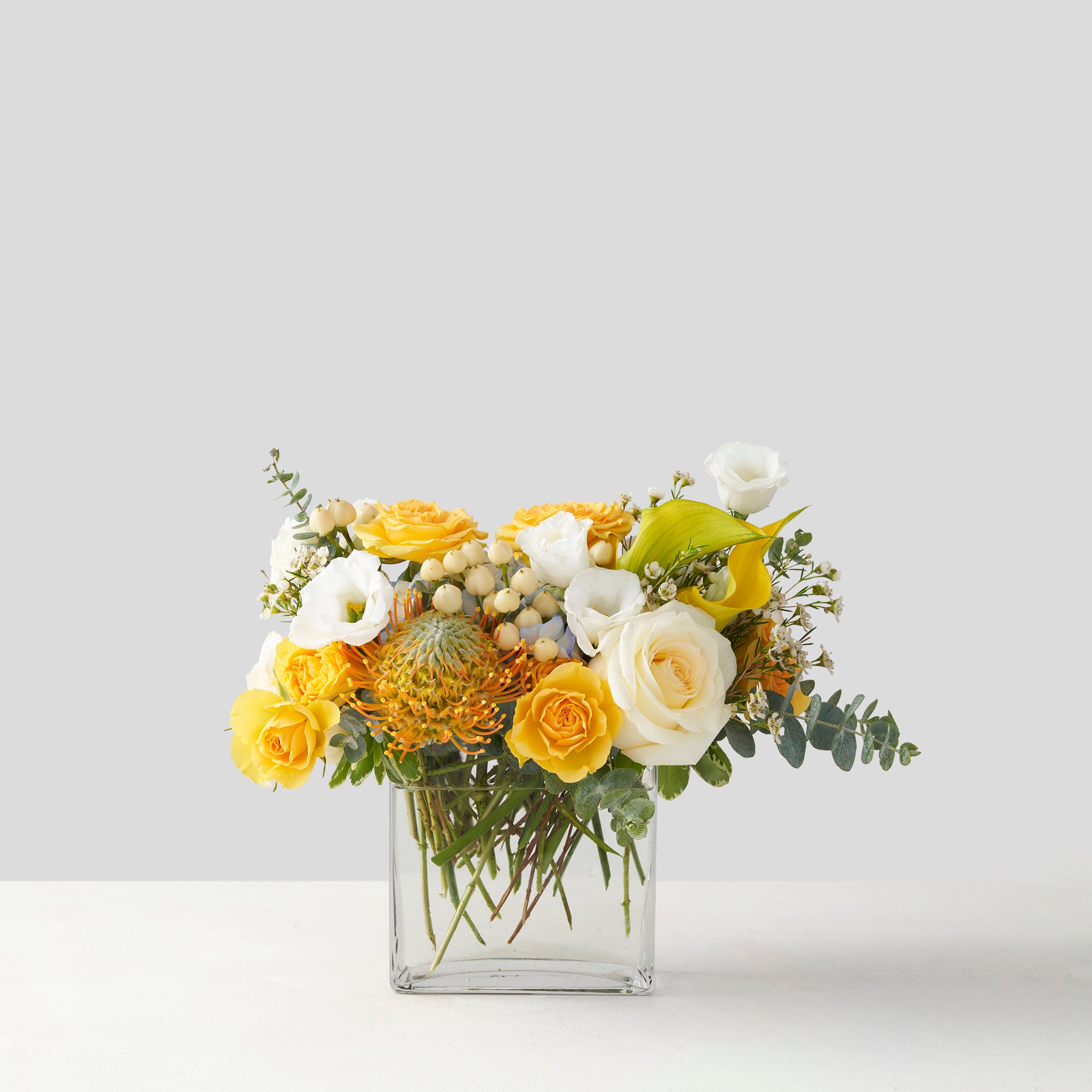 Rectangular glass vase with yellow proteal, yellow and cream roses, yellow calls, white lisianthus, and euclyptus.