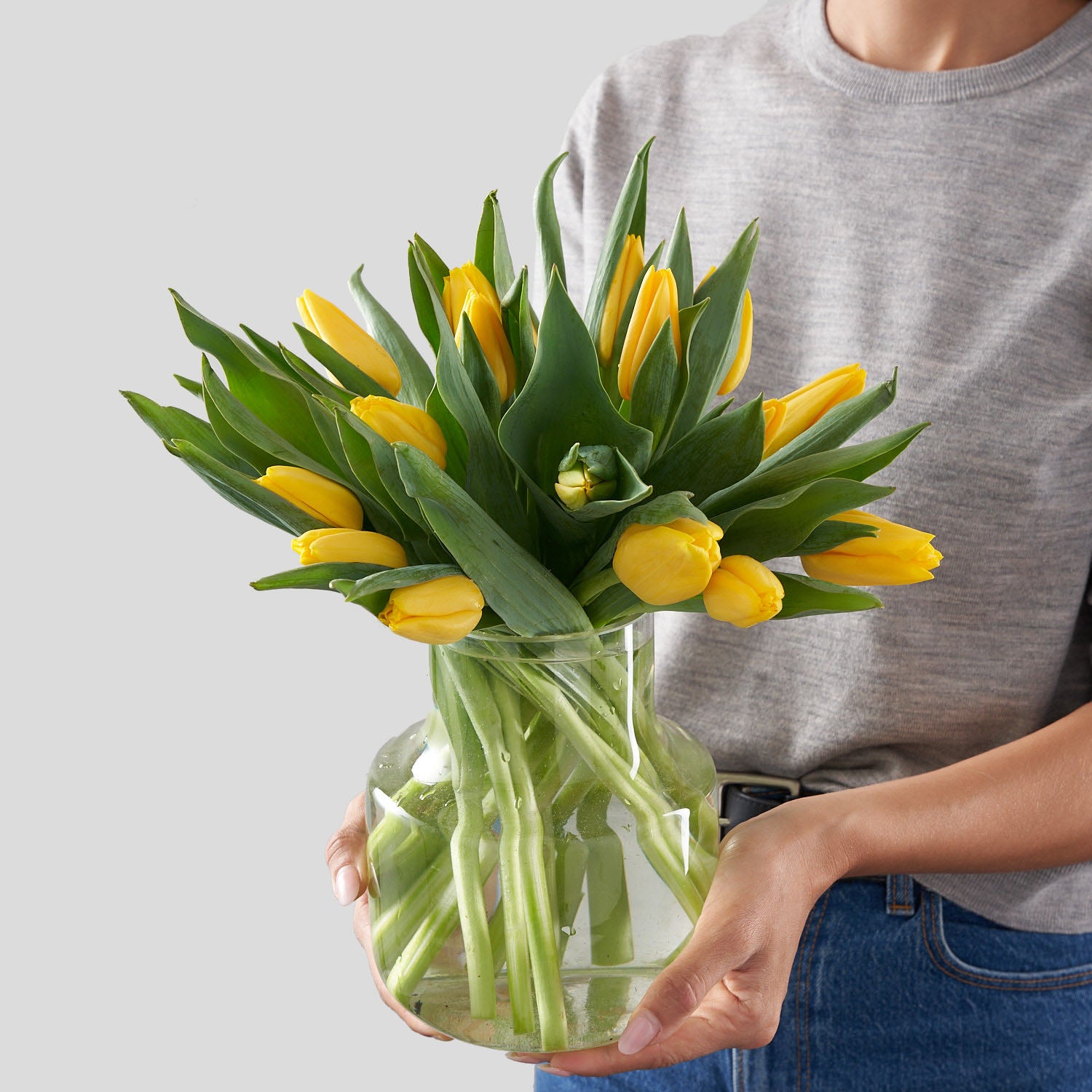 Woman in grey shirt holding a rounded clear glass vase filled with an arrangement of tulips
