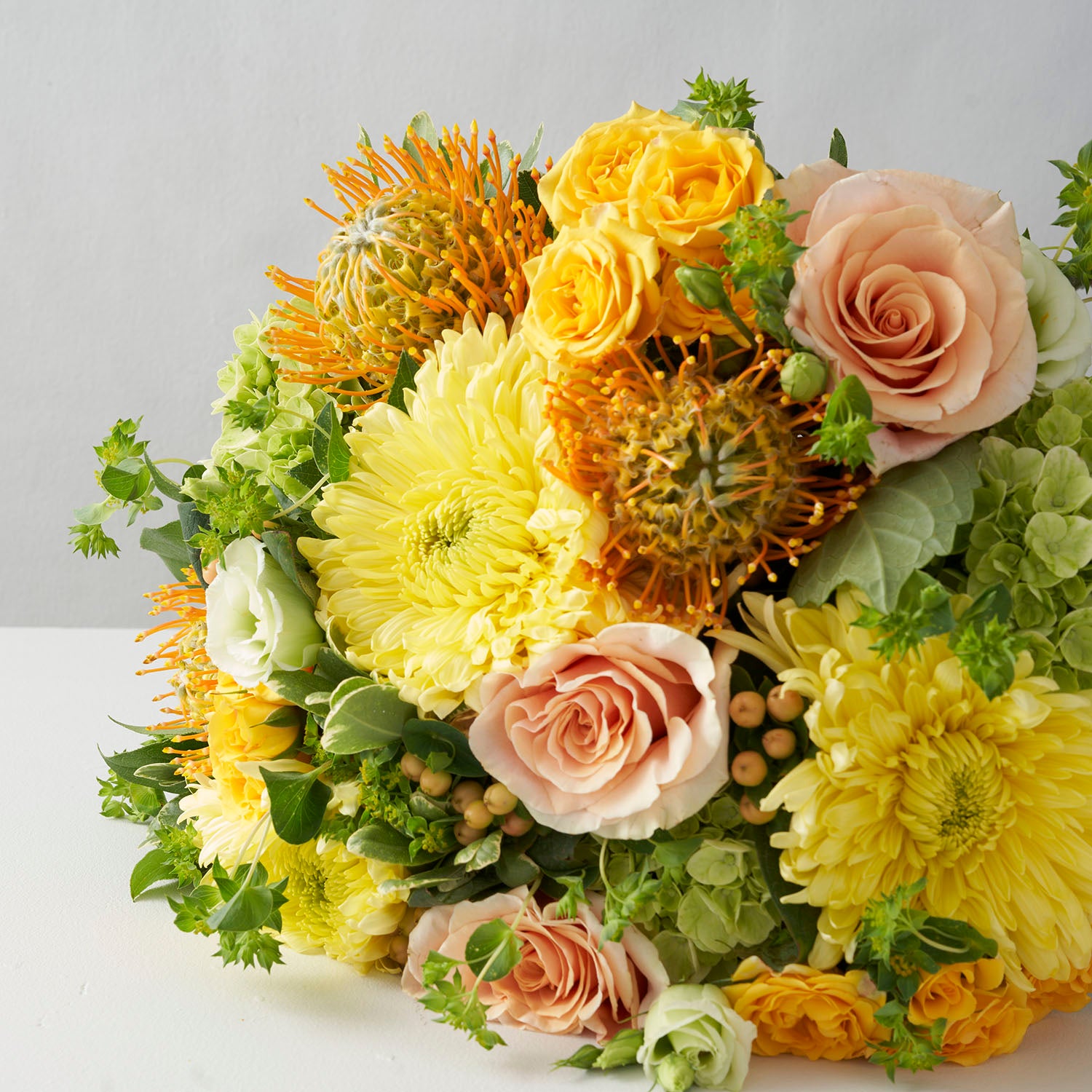 Close up of bouquet of yellow chrysanthemums, peach roses, green hydrangea, peach hypericum berries and gold protea flowers on white background.