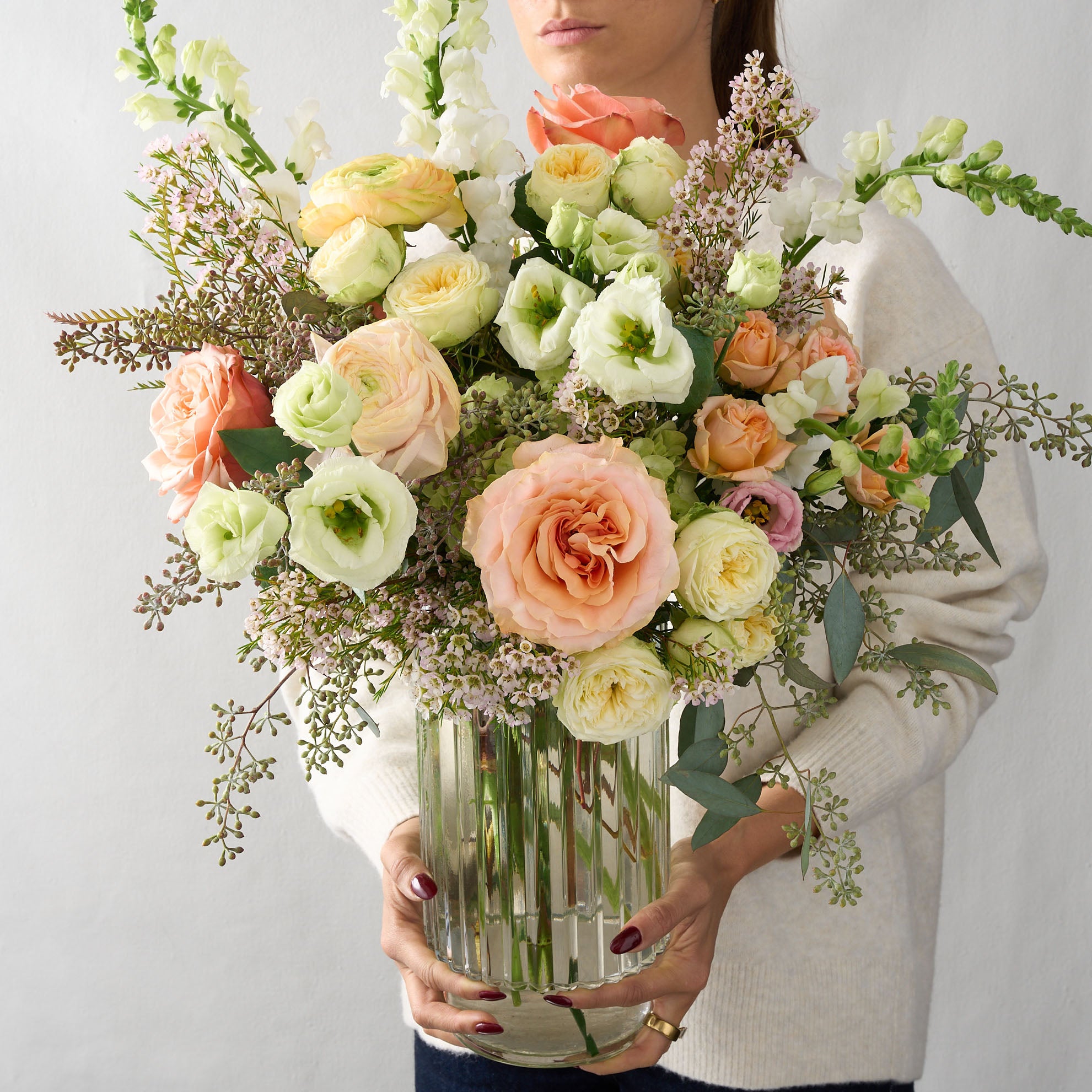 woman holding a flower arrangement white snapdragons and Lisianthus, cream double spray roses, peach roses, soft pink wax flowers and seeded eucalyptus in a clear ribbed cylinder vase