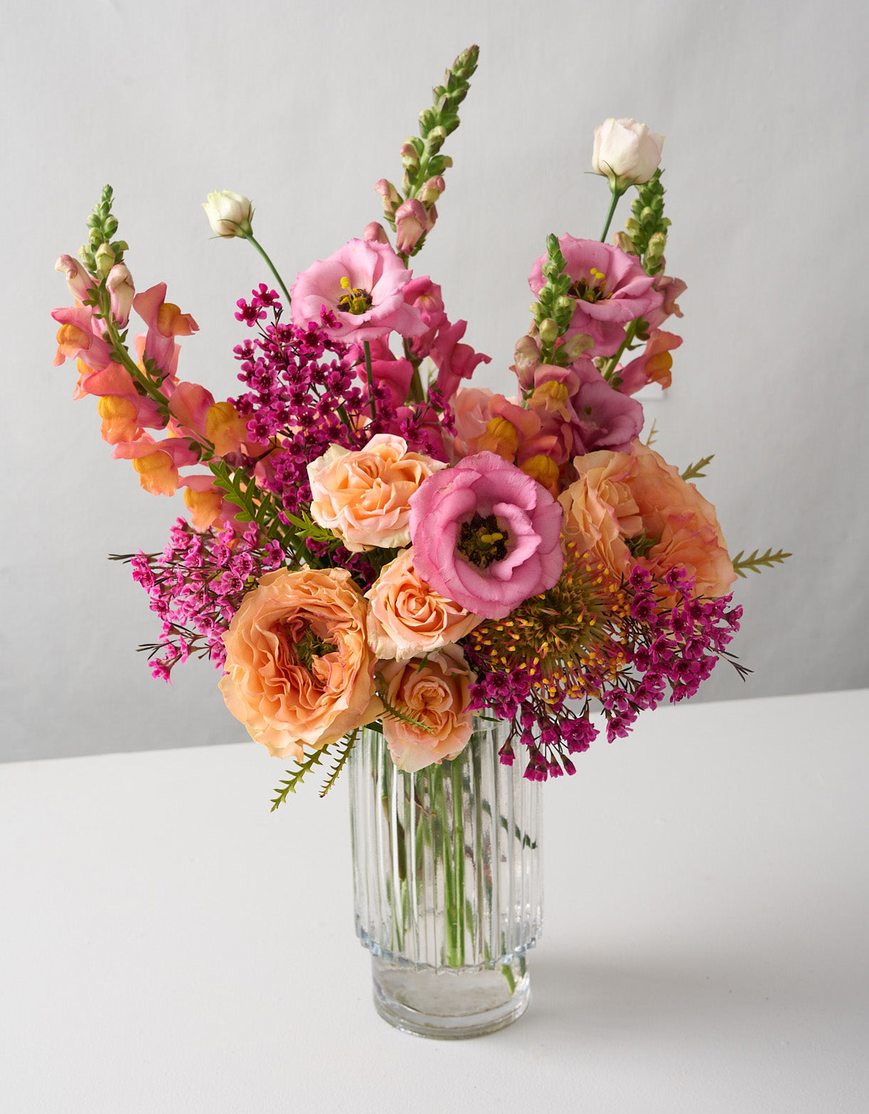 Amelia arrangement with coral snapdragons, peach roses and hot pink wax flowers in a tall cylinder beveled glass vase on a white table