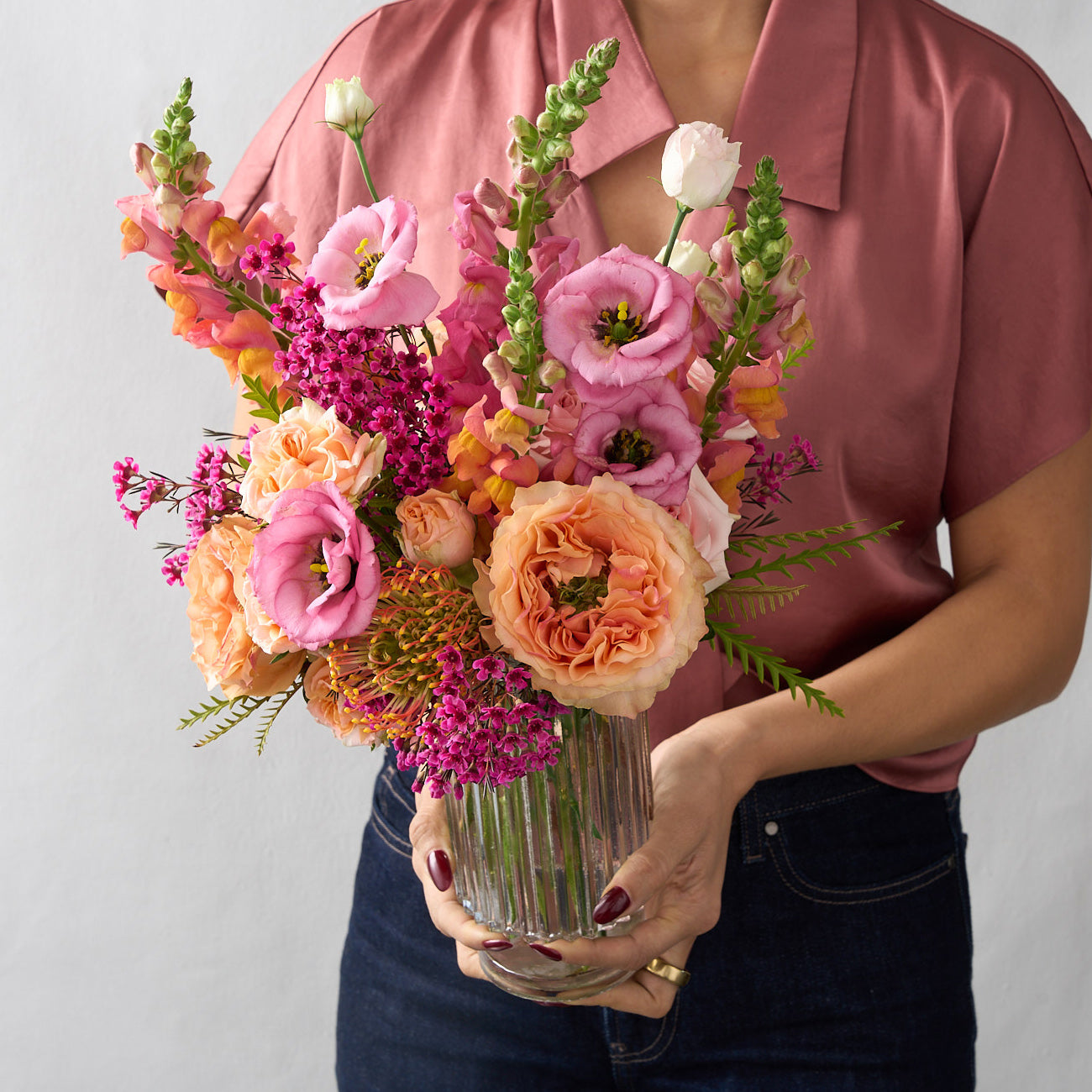 Amelia arrangement with coral snapdragons, peach roses and hot pink wax flowers in a tall cylinder beveled glass vase, held by a woman