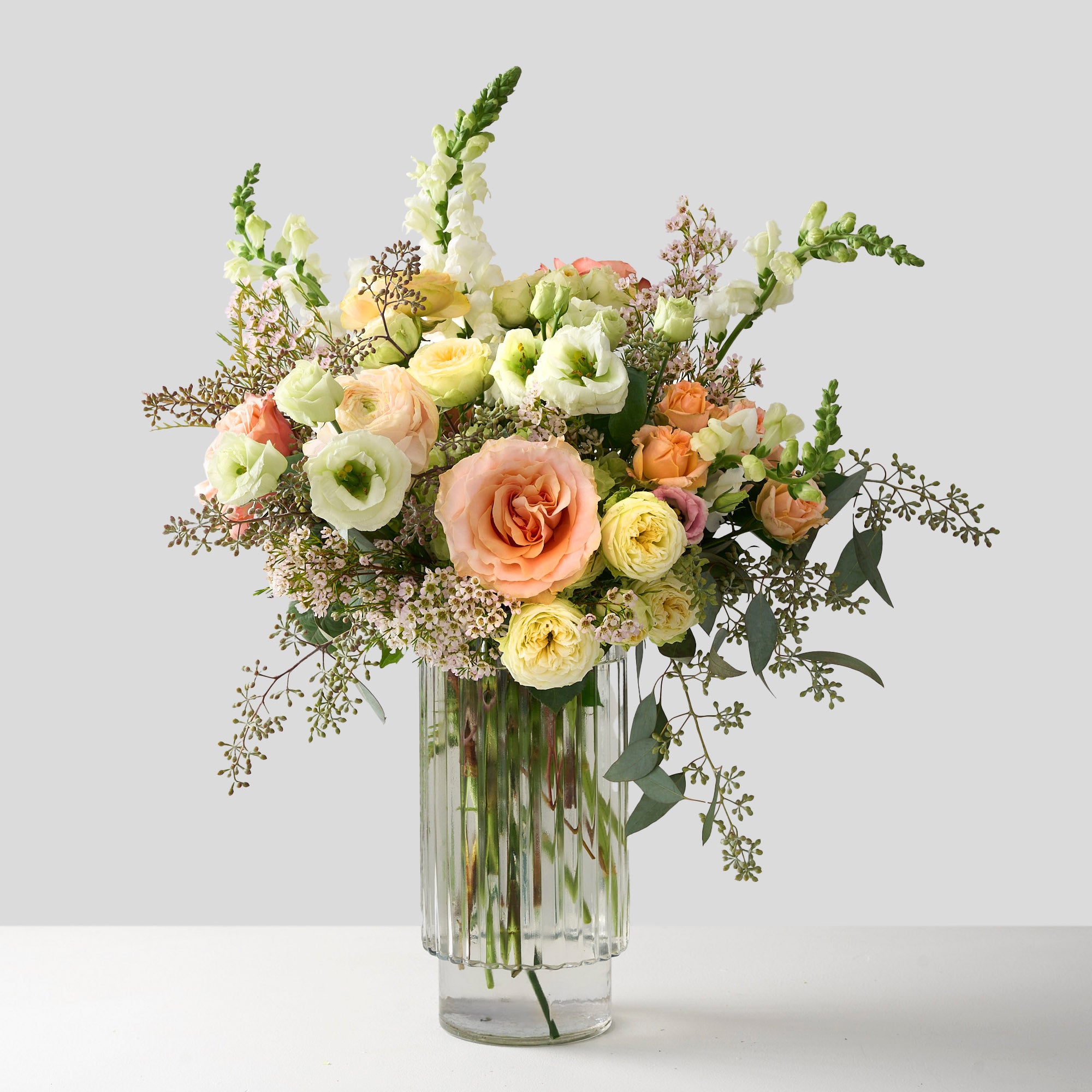 tall arrangement of white snapdragons, double spray roses, peach roses and seeded eucalyptus in a clear ribbed cylinder vase