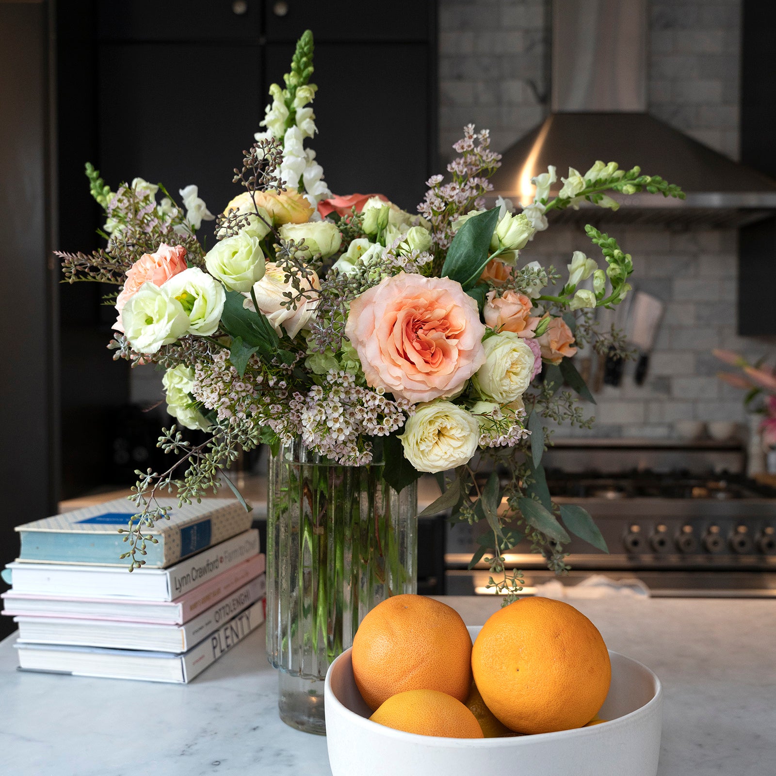 tall arrangement of white snapdragons, peach roses and seed eucalyptus on a kitchen island with an orange and books