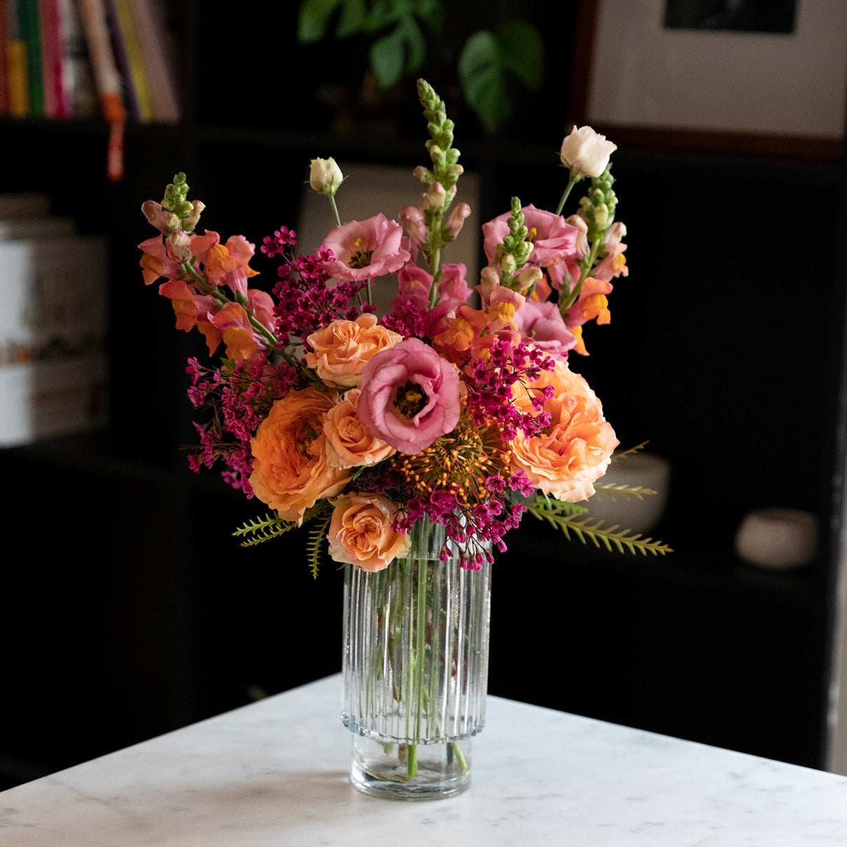 Amelia arrangement with coral snapdragons, peach roses and hot pink wax flowers in a tall cylinder beveled glass vase in front of black bookcase