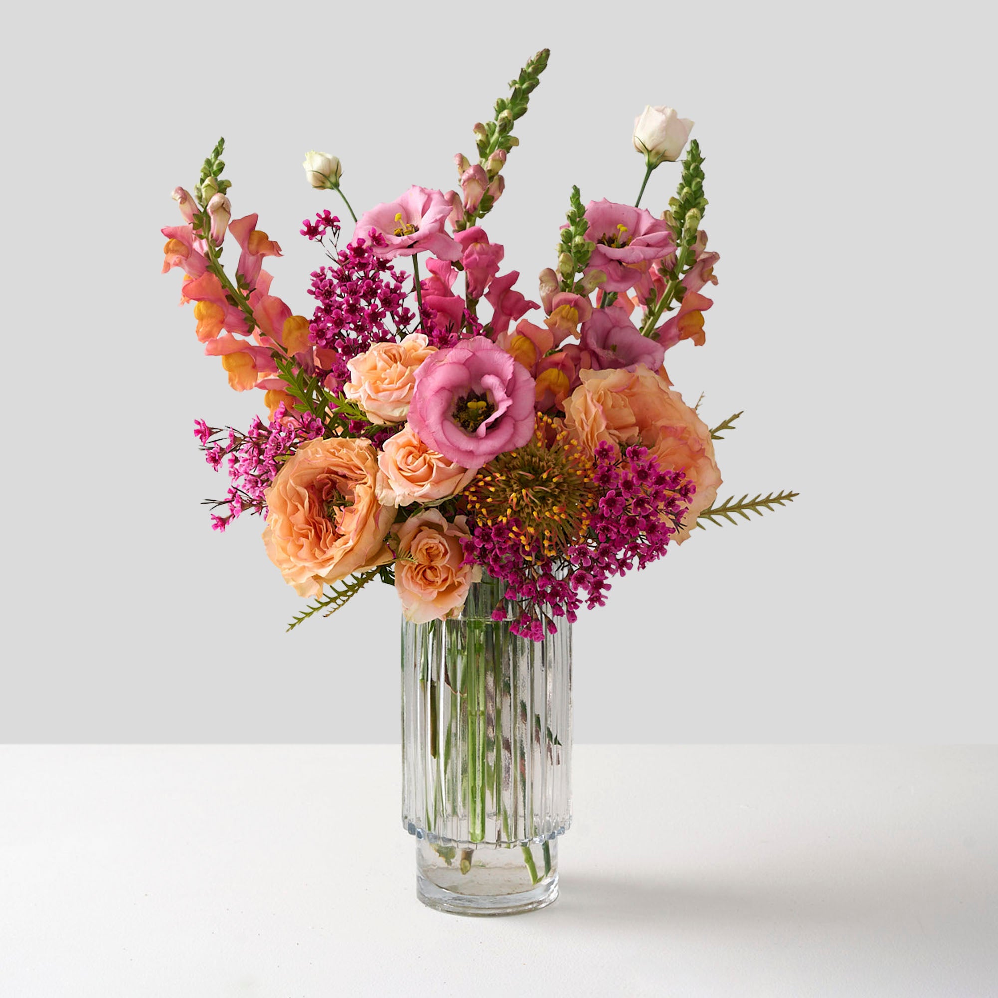 Amelia arrangement with coral snapdragons, peach roses and hot pink wax flowers in a tall cylinder beveled glass vase