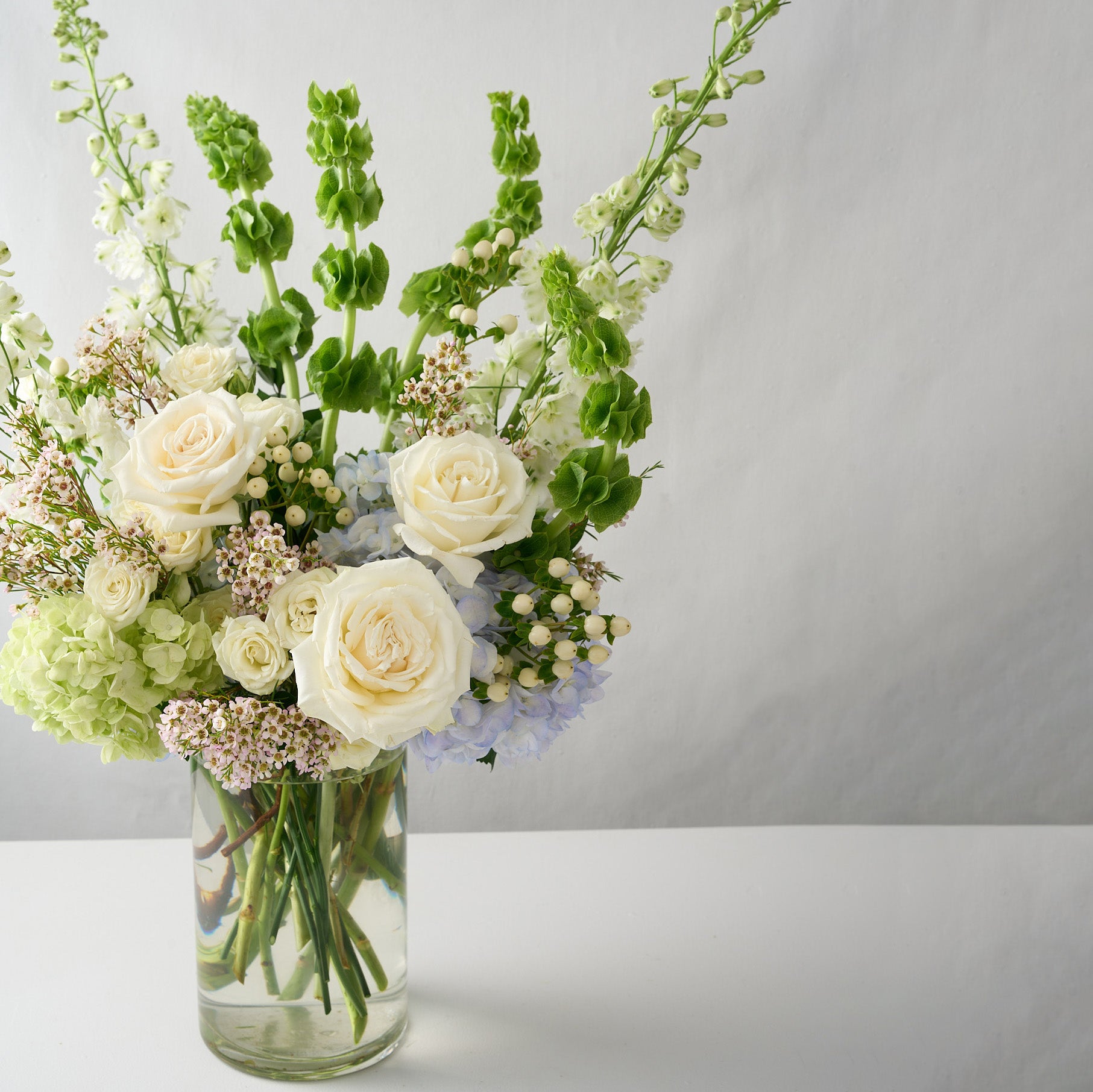 photo of a tall flower arrangement skewed to the left with white delphinium,  green bells of Ireland, blue & green hydrangea, cream candlelight roses 