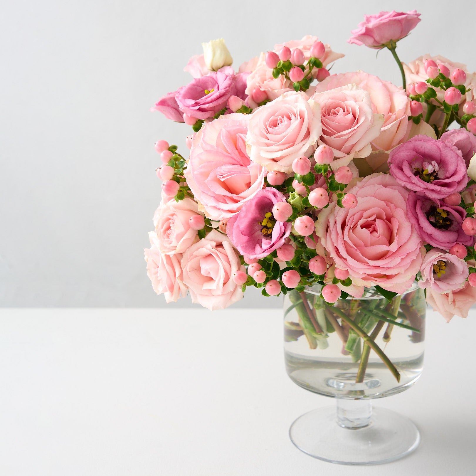 off center photo of Chloe flower arrangement with tones of pink Esperance roses, pink lisianthus, spray roses and hypericum in a clear pedestal glass vase 