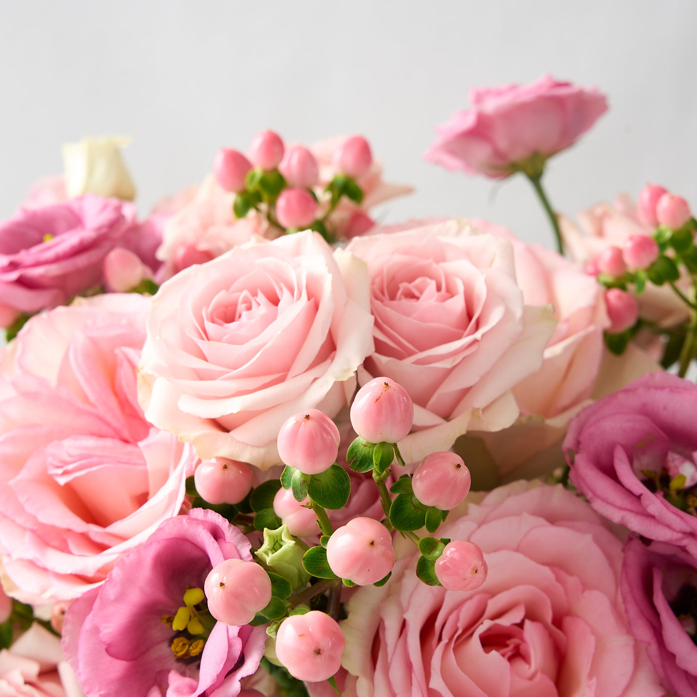 close up photo of a flower arrangement with tones of pink Esperance roses, pink lisianthus, spray roses and hypericum in a clear pedestal glass vase 