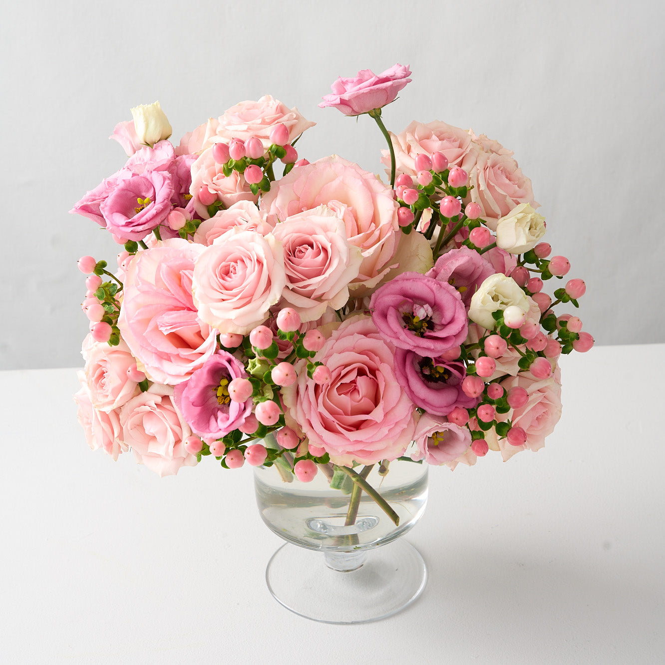 top angle photo of a flower arrangement with tones of pink Esperance roses, pink lisianthus, spray roses and hypericum in a clear pedestal glass vase 