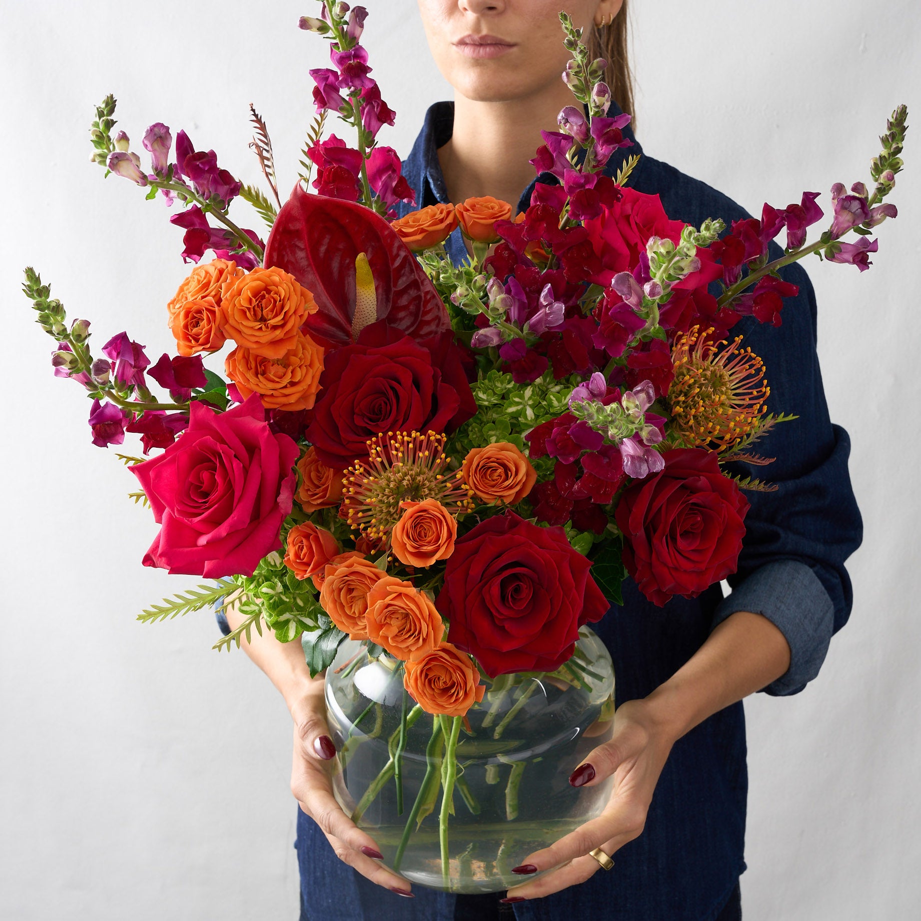 Woman holding a floral arrangement featuring deep red roses, vibrant orange hues, and striking red anthuriums in a bold and luxurious display.