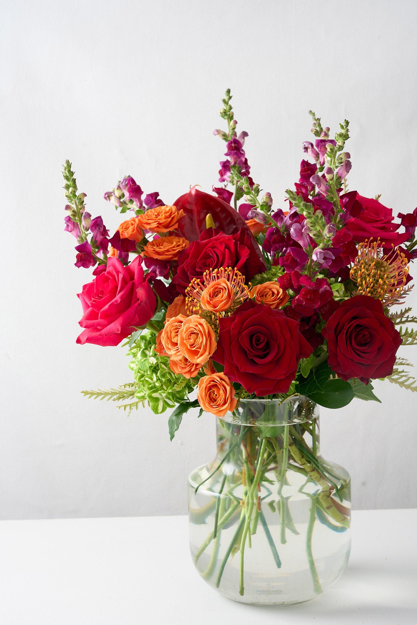 photo of a floral arrangement  skewed to the right, featuring deep red roses, vibrant orange hues, and striking red anthuriums in a bold and luxurious display.