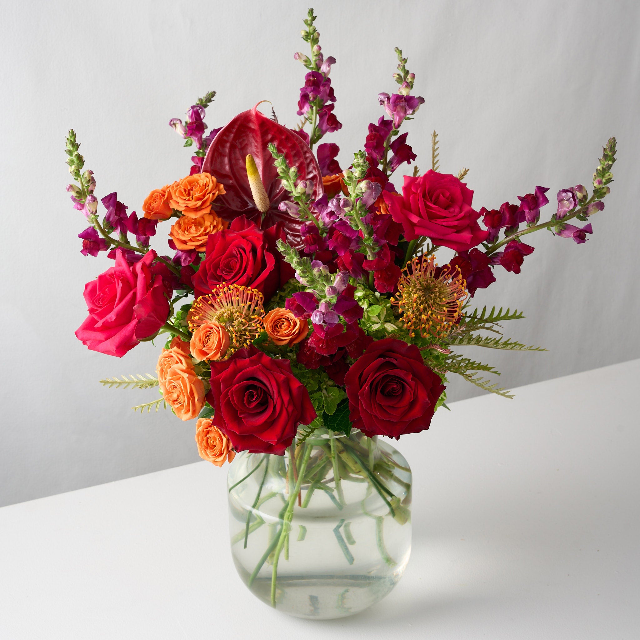 artistic photo of a floral arrangement featuring deep red roses, vibrant orange hues, and striking red anthuriums in a bold and luxurious display.