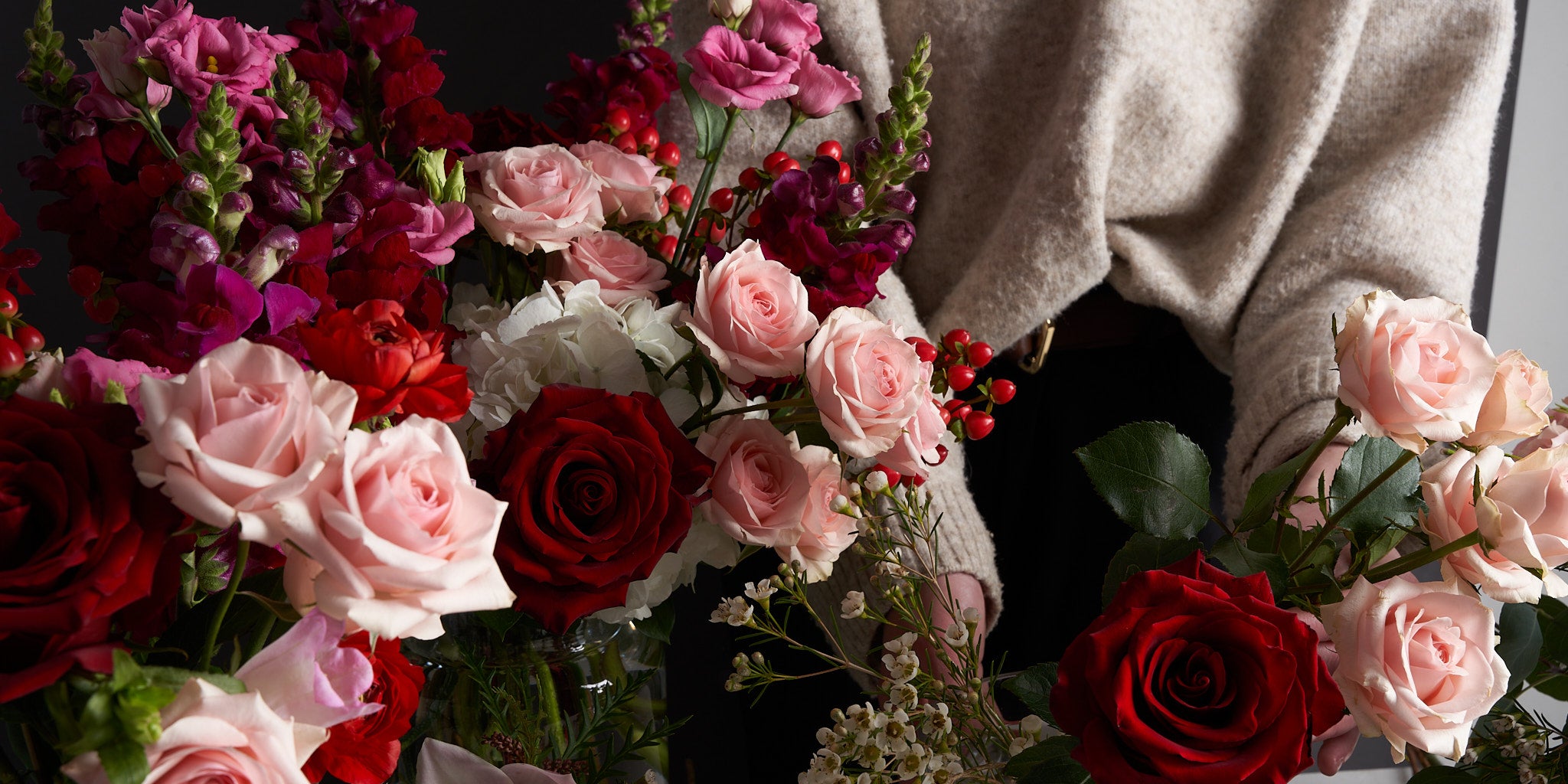 Woman in grey sweater arranging various coloured roses and anemones, with baby's breath and red hypericum berries