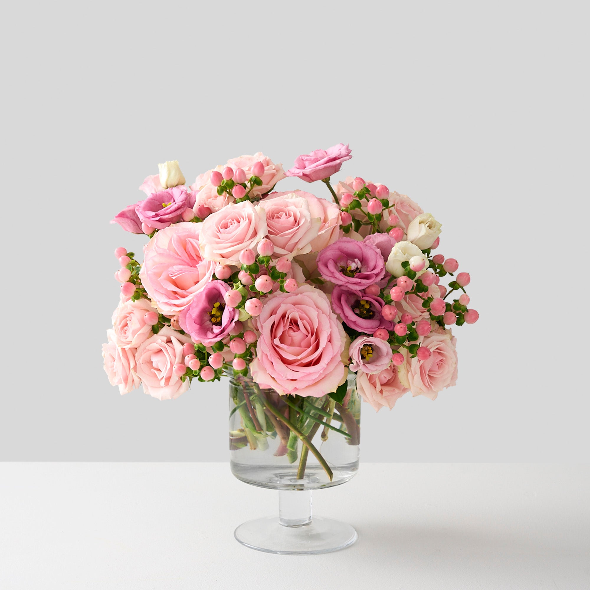  flower arrangement with tones of pink Esperance roses, pink lisianthus, spray roses and hypericum in a clear pedestal glass vase 