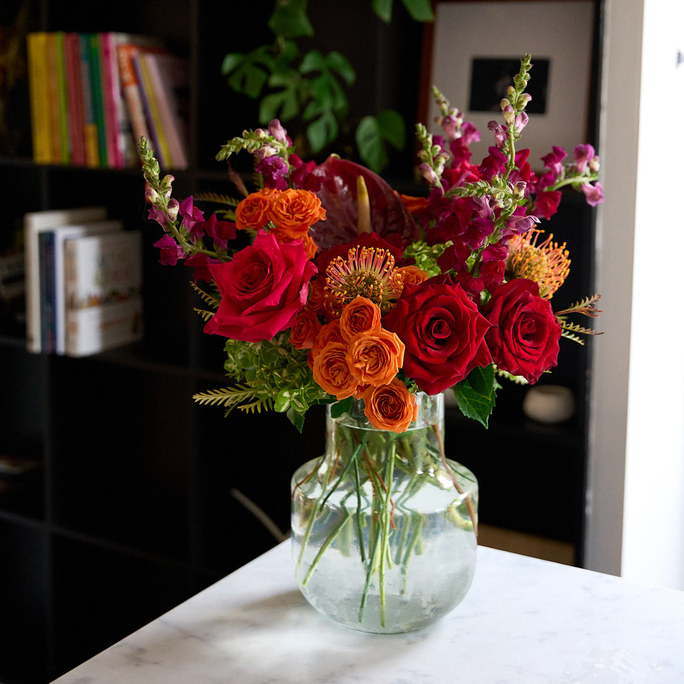 clear mild jug vase arrangement, with protea, anthurium,   red roses, snapdragons and orange spray roses