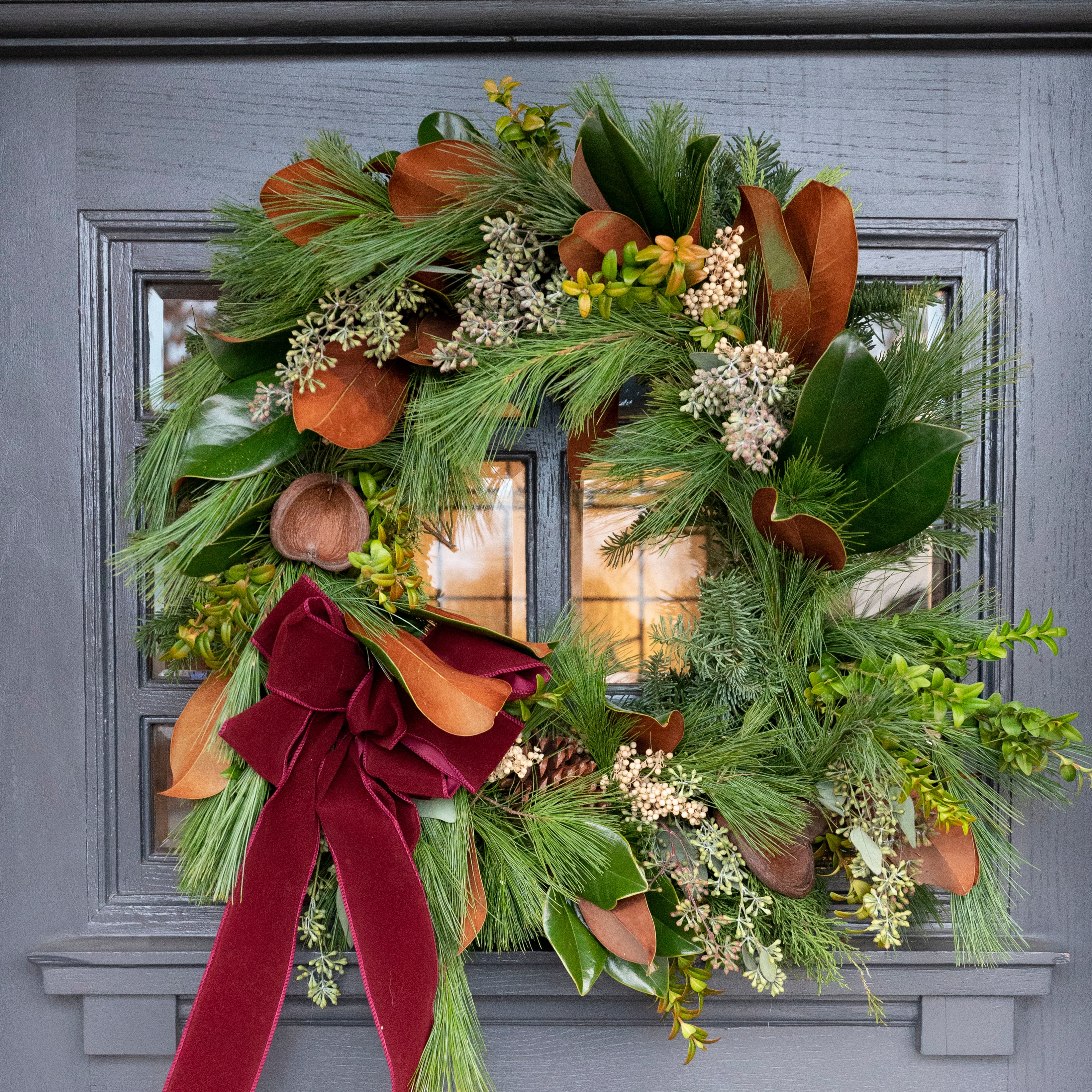 Fresh Christmas wreath with pepper berries, badam pods, magnolia leaves, cedar, fir and pine greens with a burgundy bow on a dark grey wooden door.