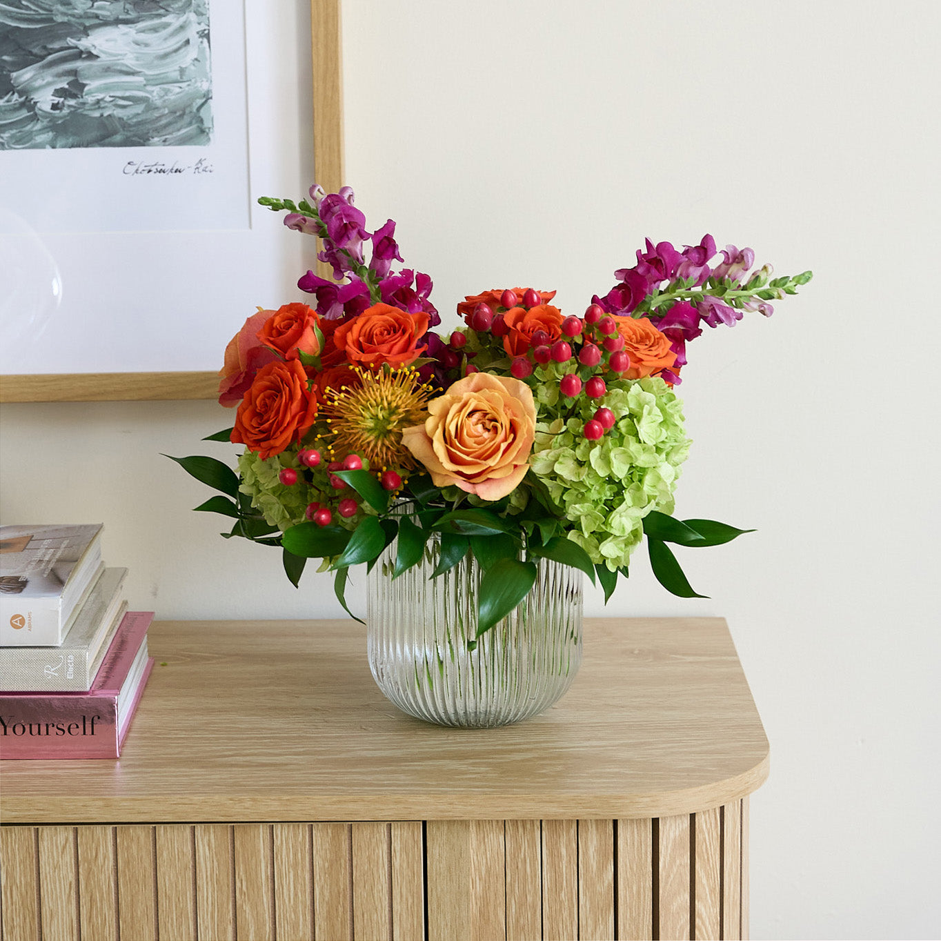coral roses and hypericum, green hydrangea , orange spray roses, fuchsia snapdragons, and protea in a clear ribbed short cylinder  vase  on a credenza with books