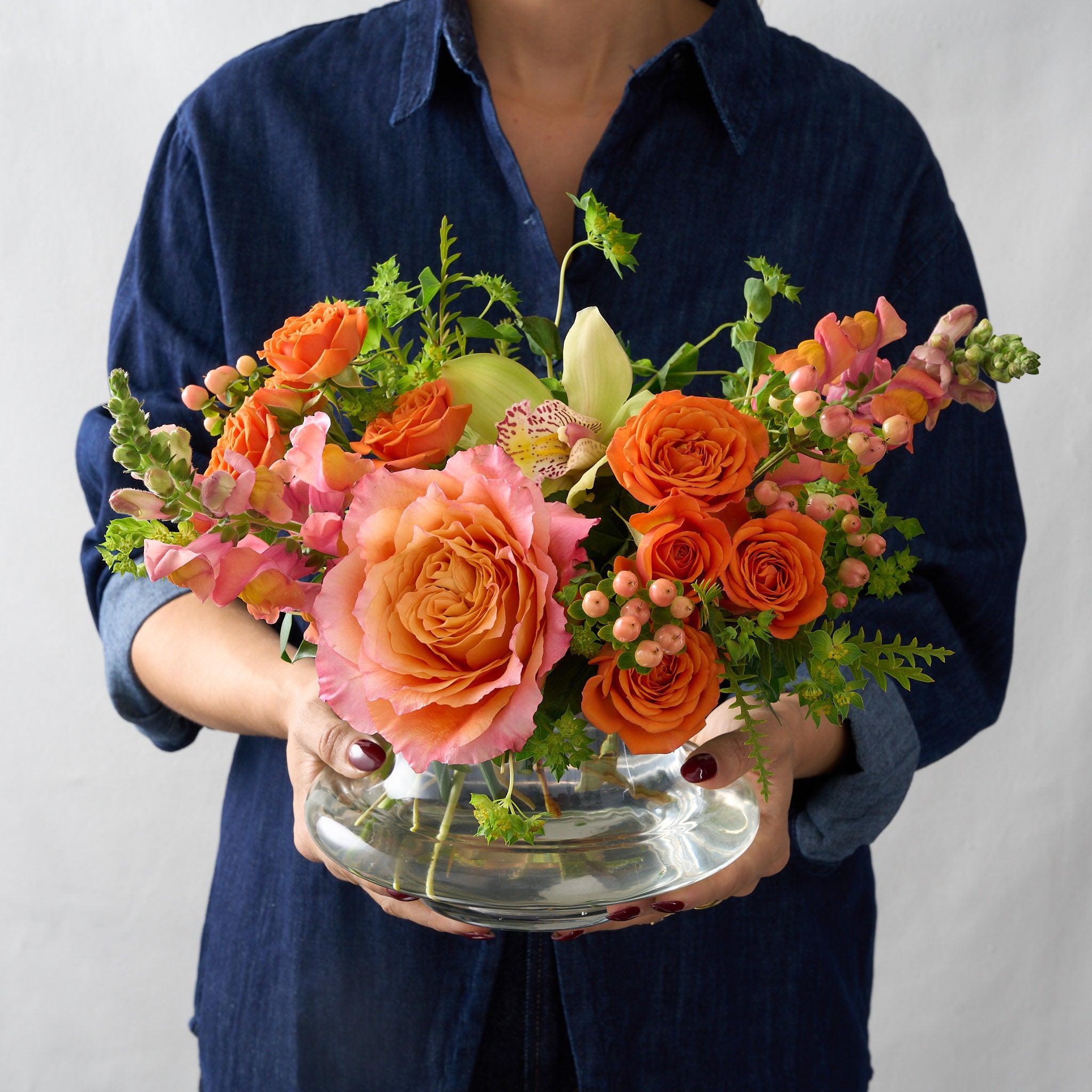 ellie flower arrangement in clear glass vase with orange spray roses, peach snapdragons, green cymbidium orchid and Bupleurum, held by a woman wearing a blue Demin shirt