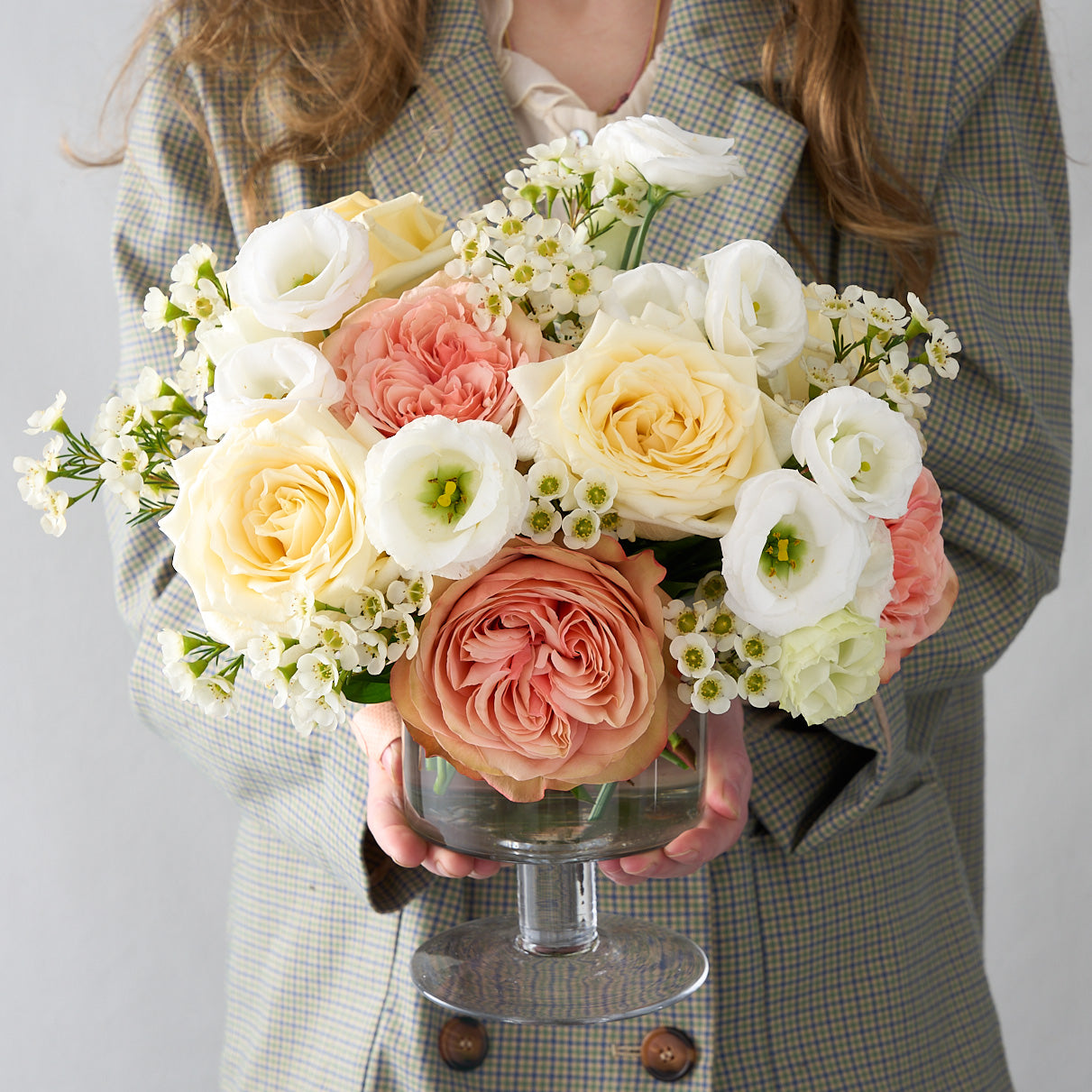 woman holding  an arrangement of pink khala  and creamy  white candlelight roses, white wax flowers and lisanthus in a pedestal glass vase.