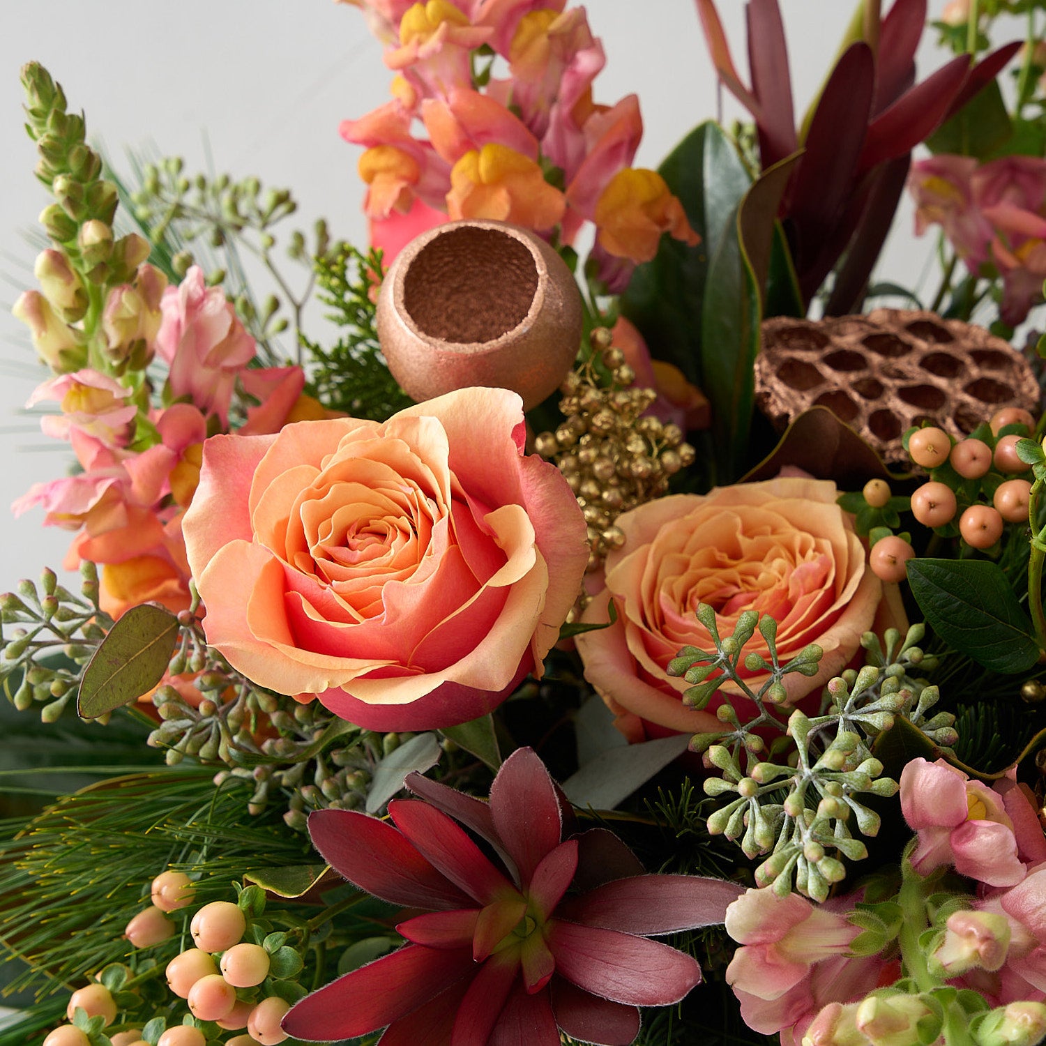 close up picture of a floral arrangement with peach toned snapdragons, roses and hypericum, gold coloured lotus pods, seeded eucalyptus and pine branches in a gold pot.