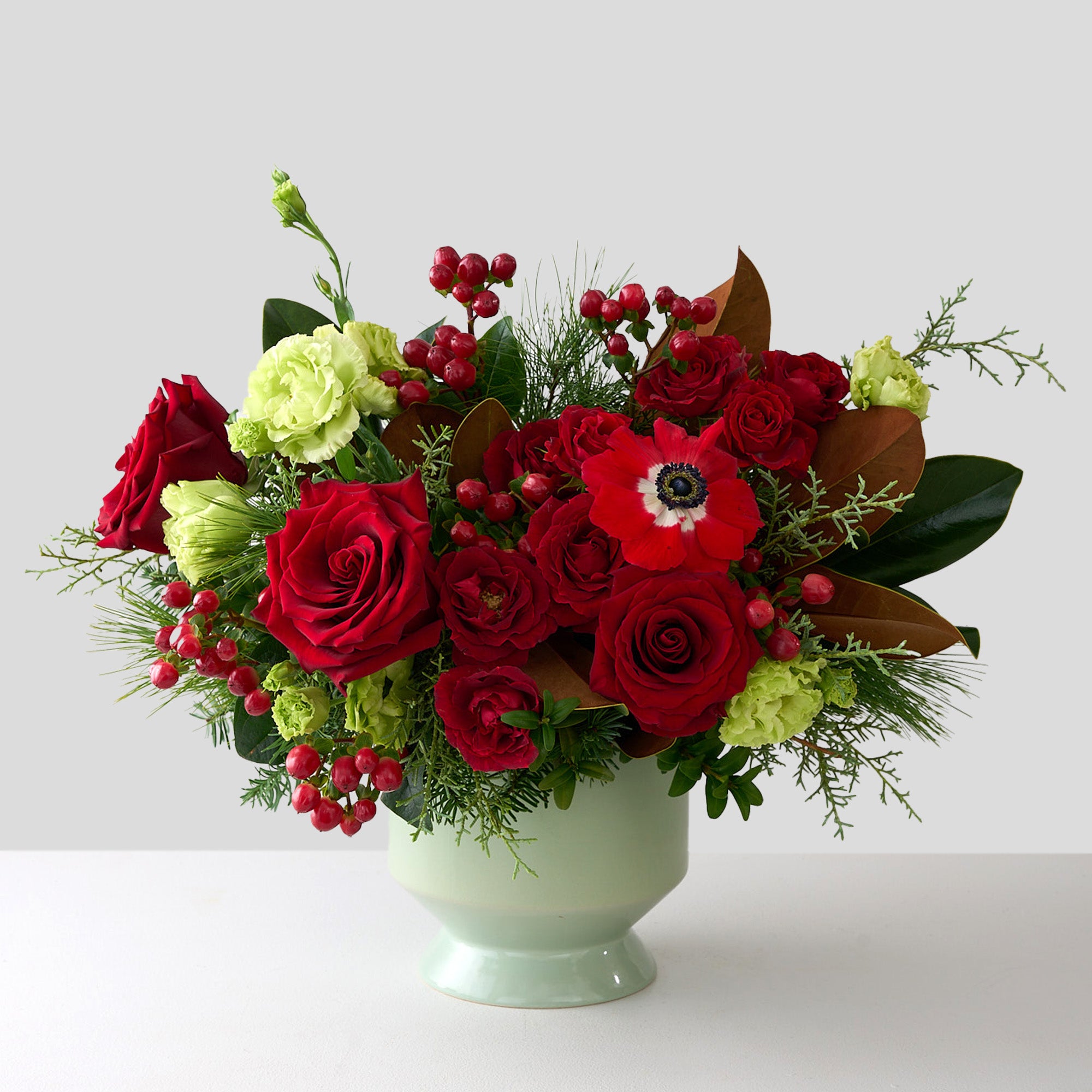 Christmas arrangement with double green Lisianthus, red roses, hypericum, and winter greens in a sage pedestal ceramic vase.