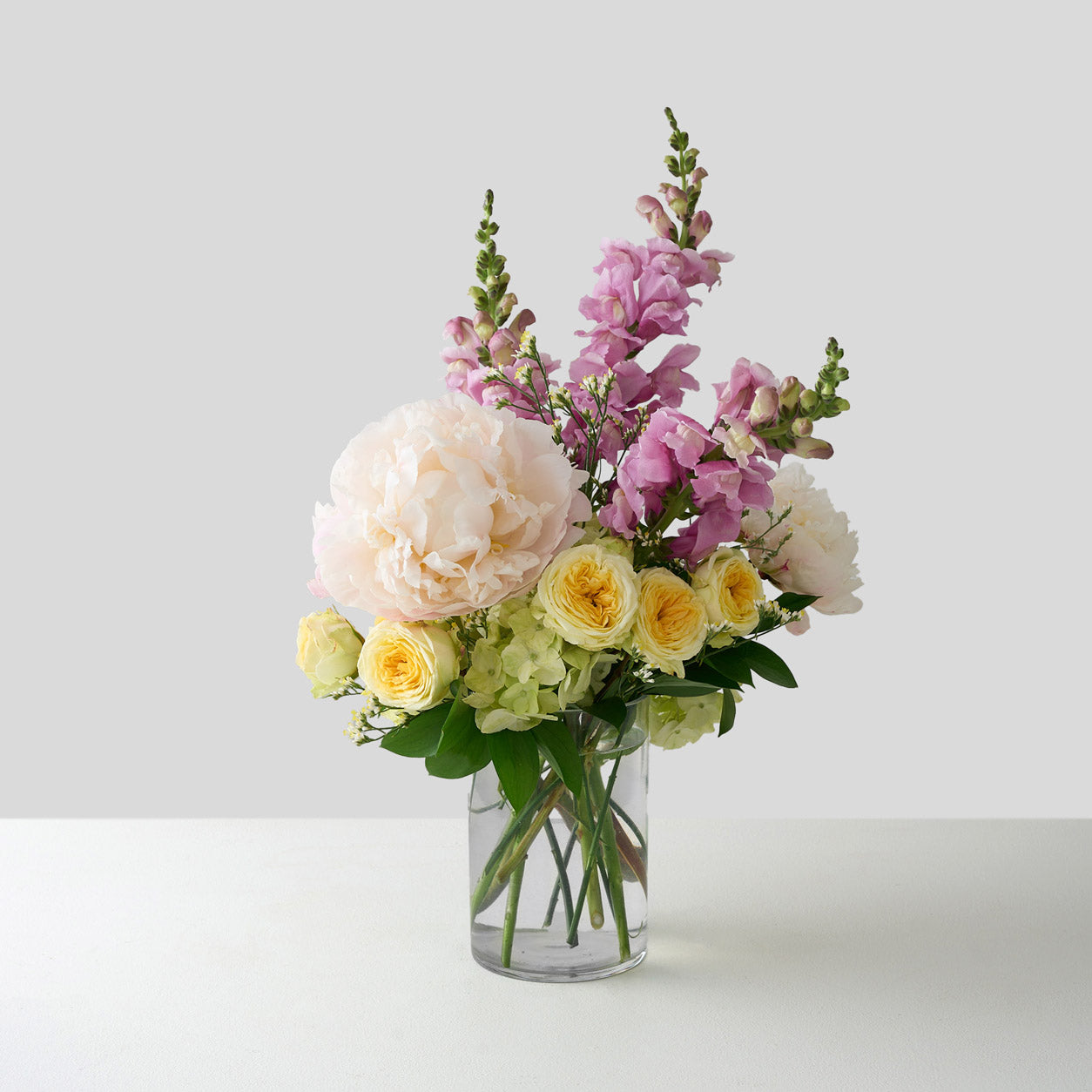 two white peonies with yellow spray roses, pink snapdragons and white hydrangea in a small clear vase.