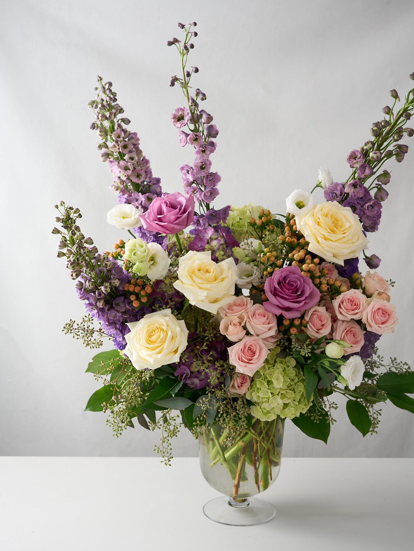 off center photo of a stunning arrangement showcases tall purple delphiniums and hydrangeas, paired with bold Candlelight roses and spray roses, while delicate seeded eucalyptus in a large glass pedestal  vase